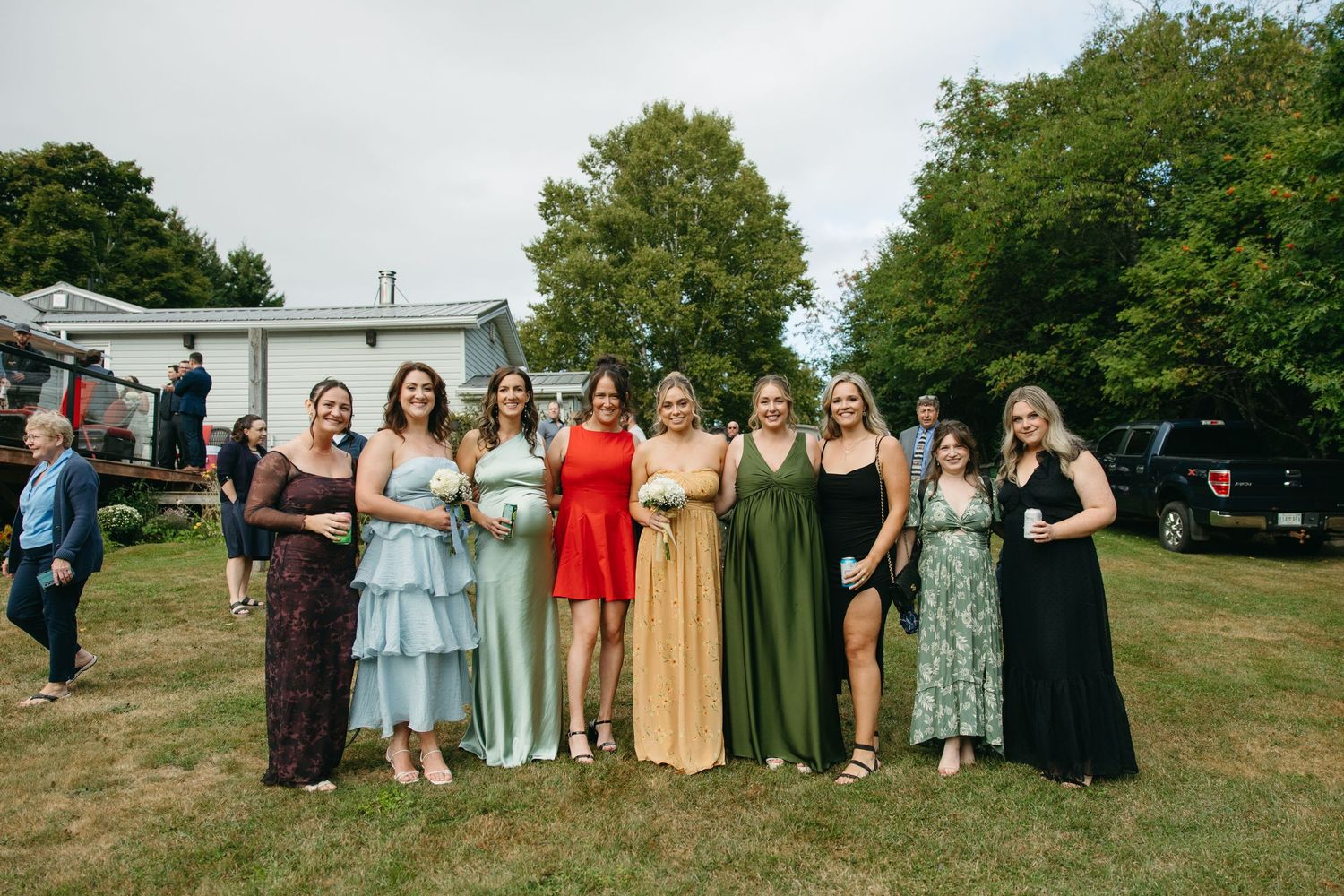 Group photo of wedding attendees wearing coordinated dresses in various colors standing on grass.