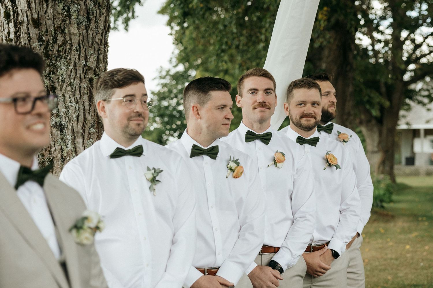 Groomsmen in white shirts and green bow ties with boutonnieres stand together at outdoor wedding.