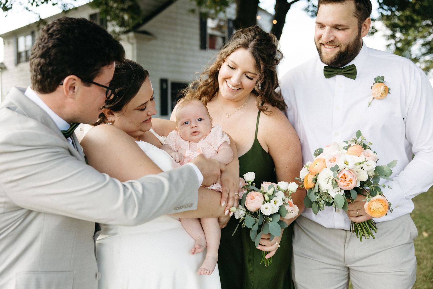 Wedding party smiles at adorable baby in pink outfit outdoors, bride in white, bridesmaid in green dress holding peach bouquet.