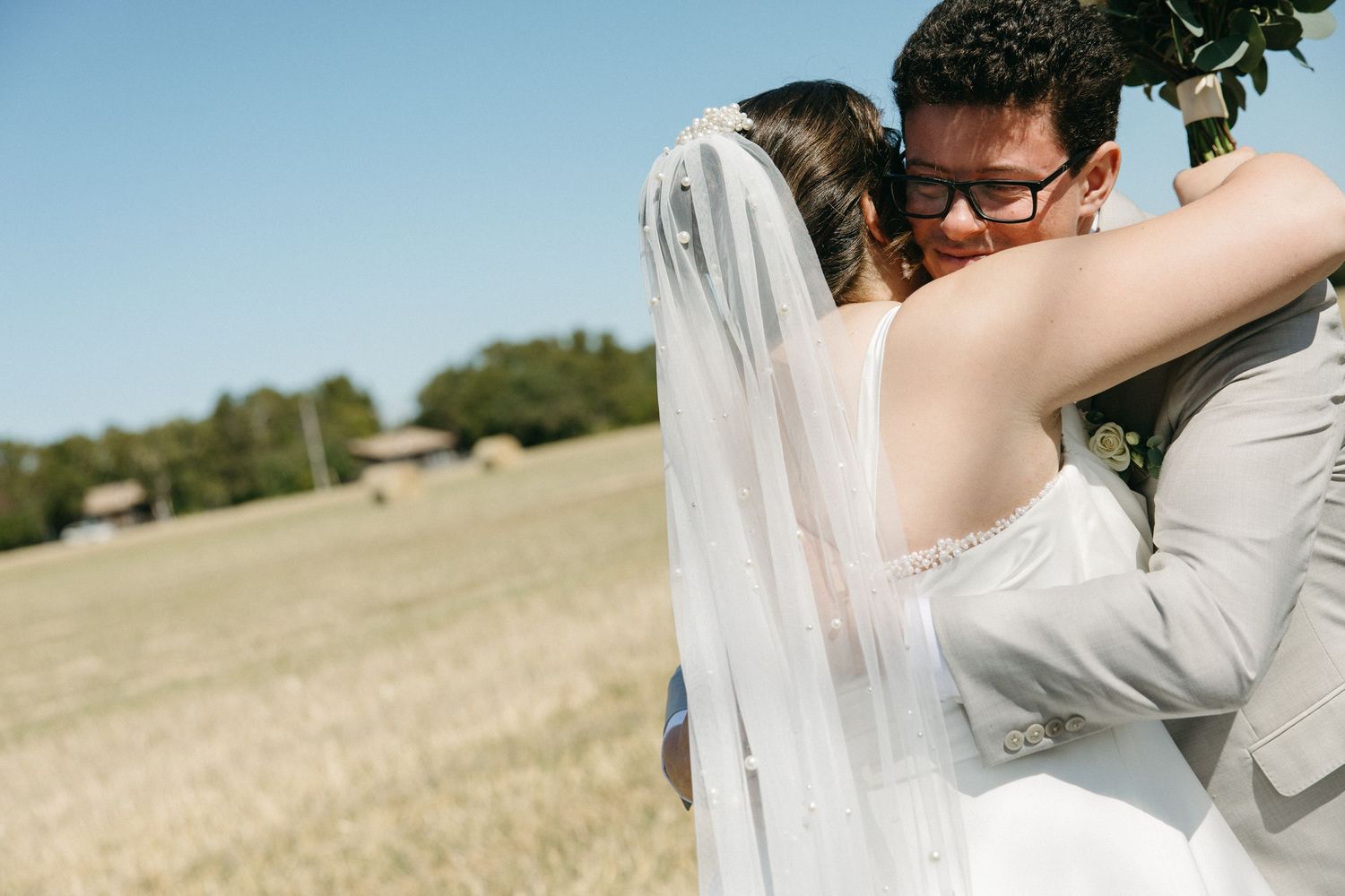 A bride and groom share a tender embrace in an open golden field on a sunny wedding day.