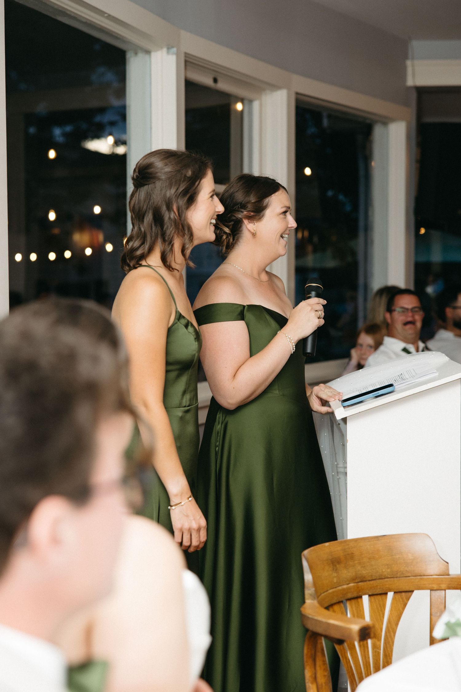 Two bridesmaids in elegant green satin gowns deliver a heartfelt speech at a wedding reception with string lights.