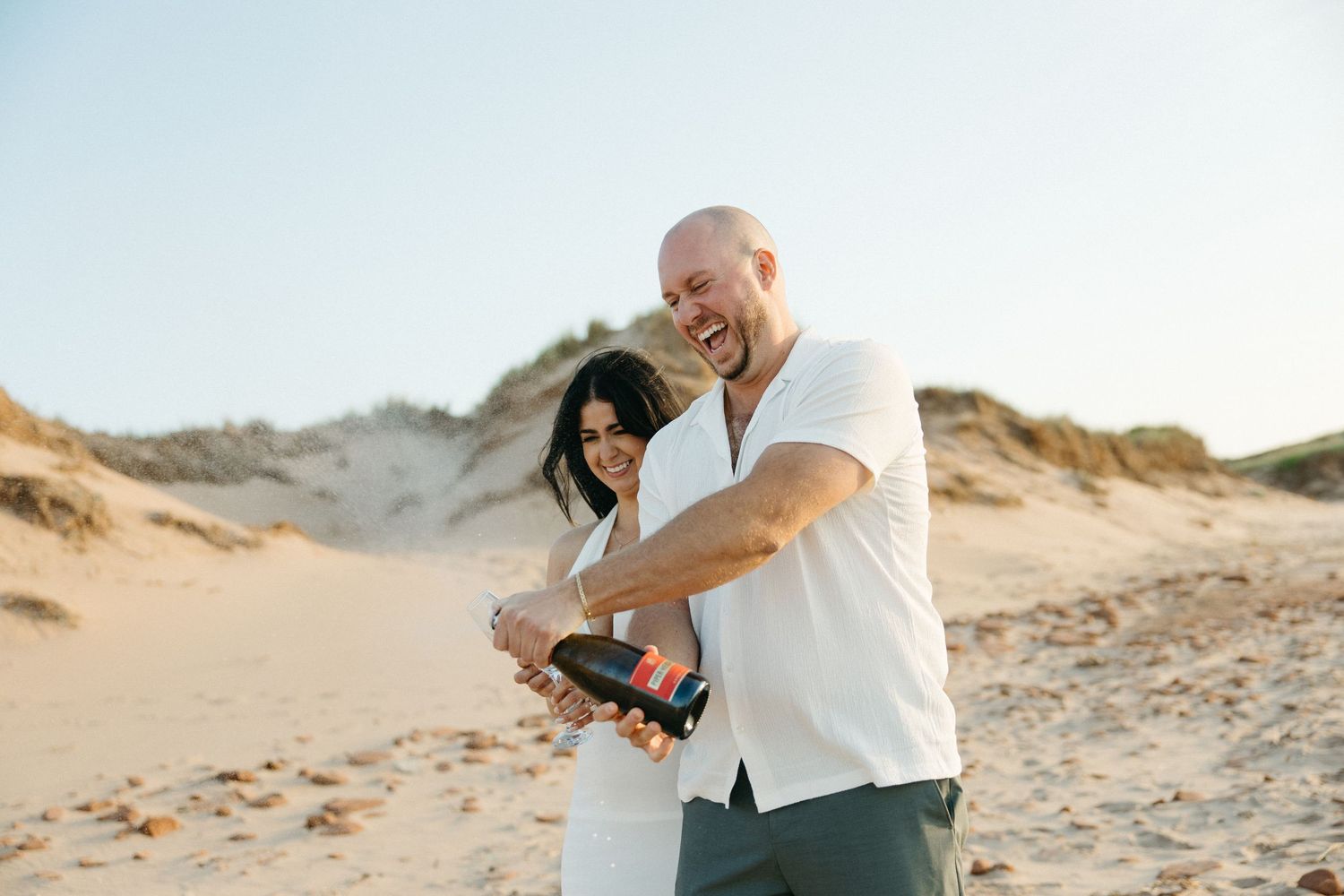 Joyful couple popping champagne bottle together on sandy beach dunes at golden hour.