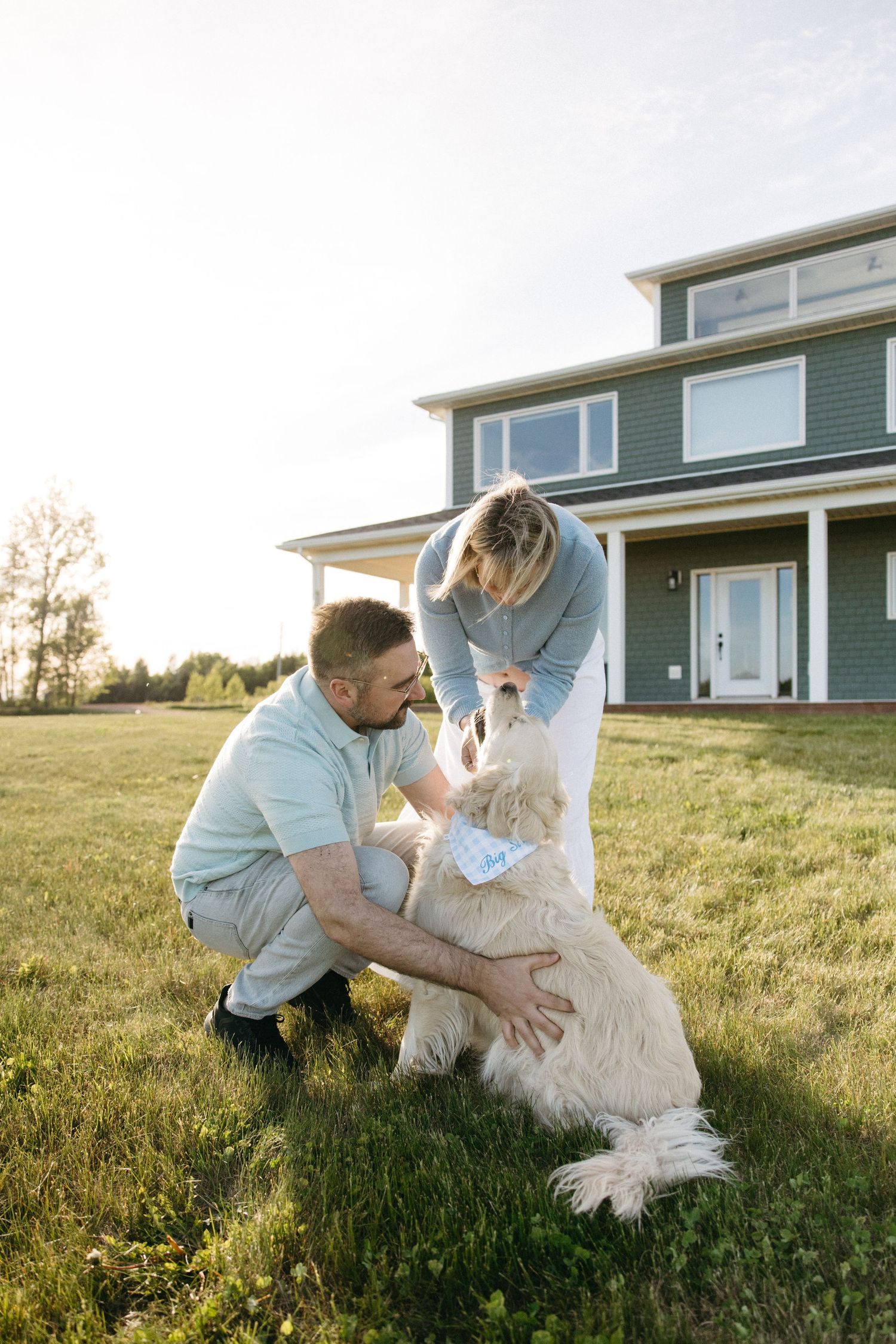 Man and woman petting a large white dog on grass in front of a modern green two-story home.