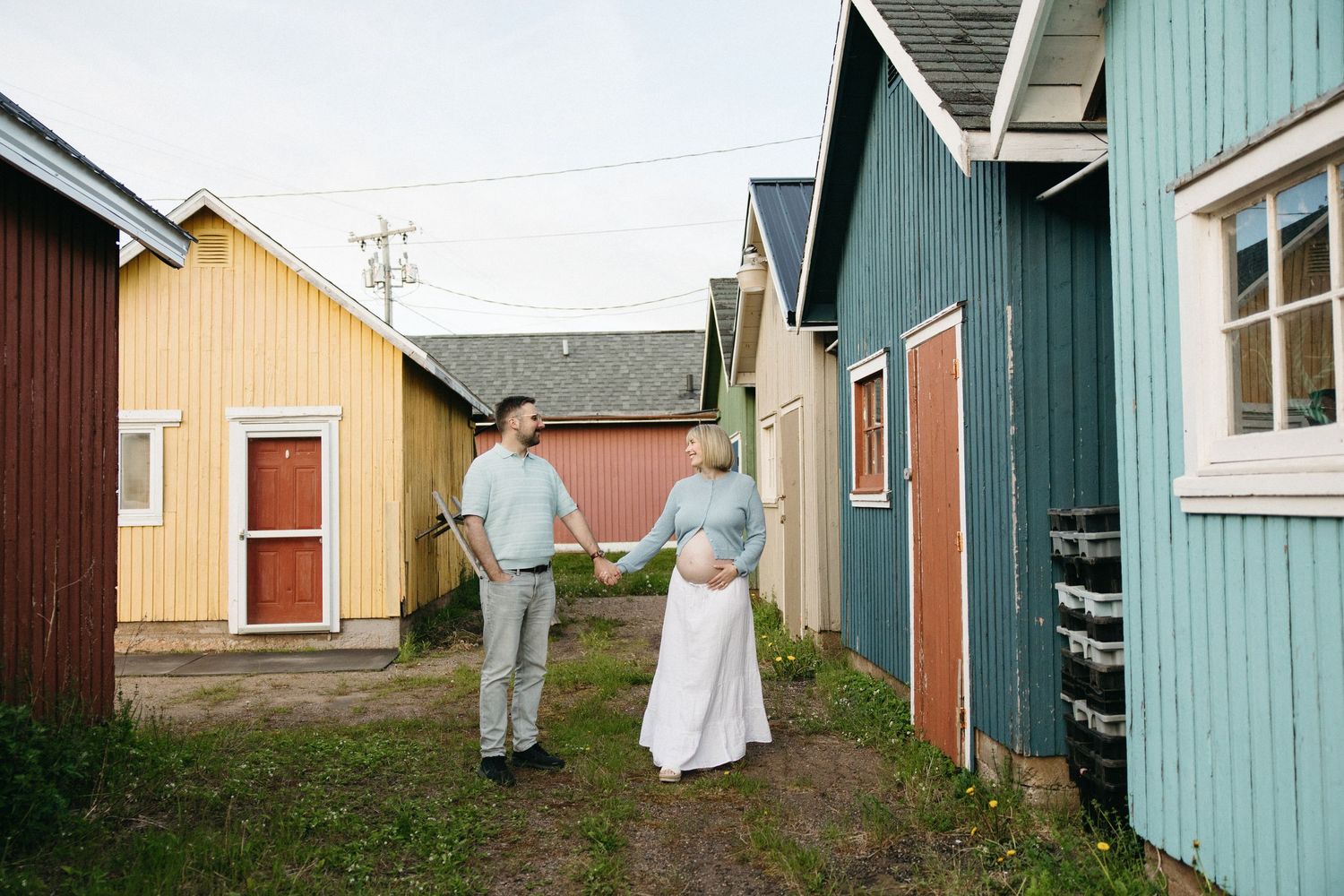 Pregnant couple holding hands between colorful wooden boathouses during an outdoor maternity photo session.
