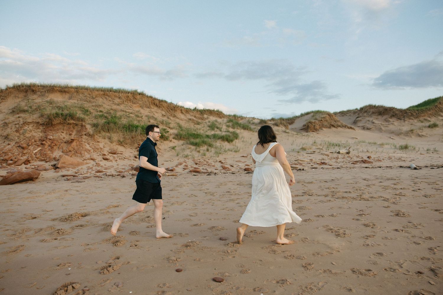 Couple walking barefoot on a sandy beach with grass-covered dunes in the background.