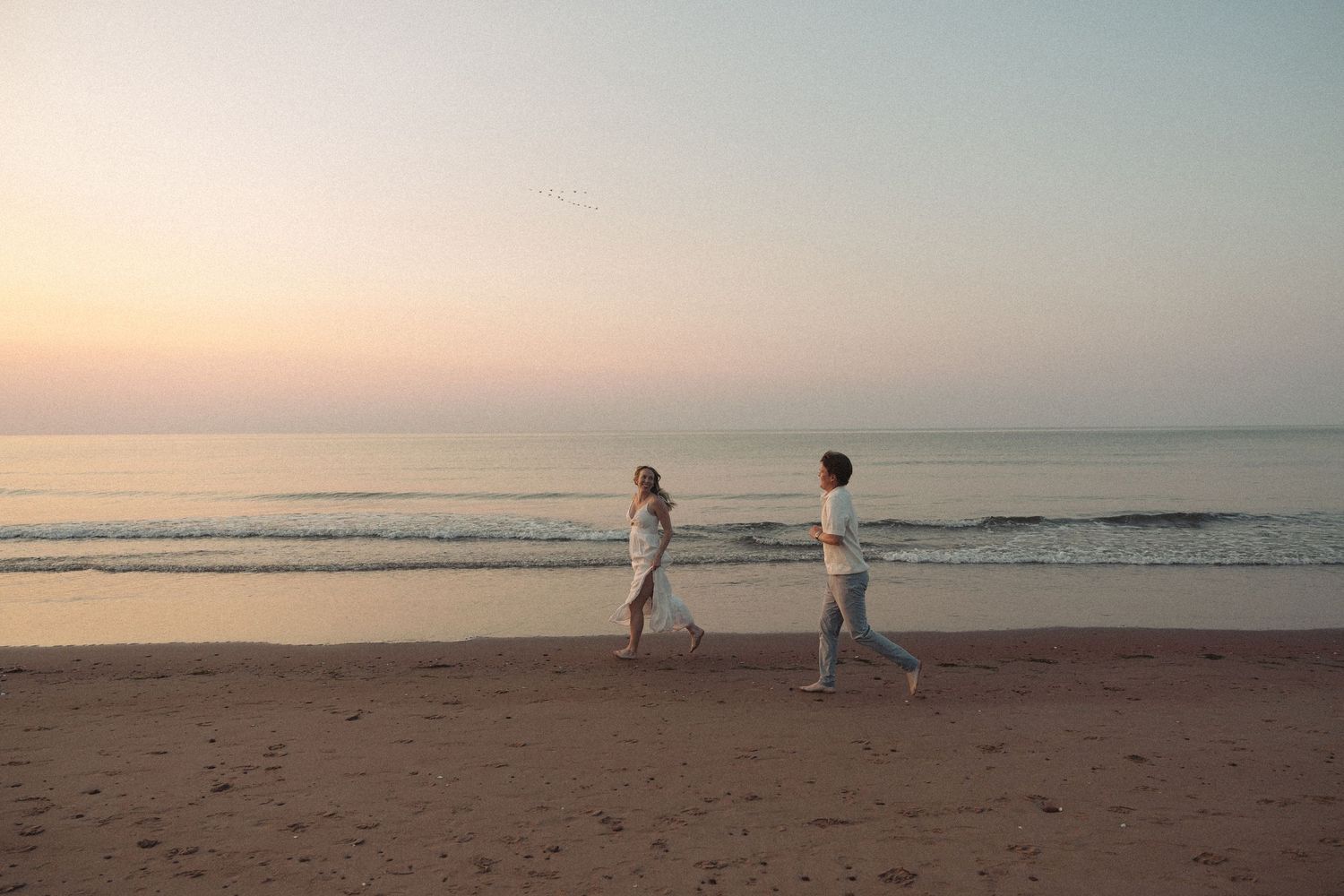 Couple walking along sandy beach at sunset with calm ocean and pastel sky in background.
