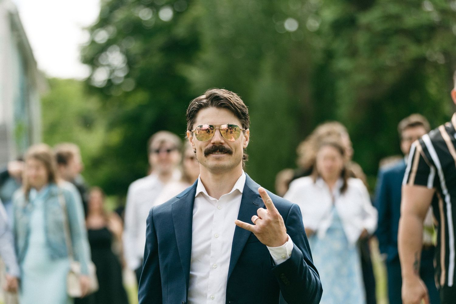 Man with mustache and sunglasses in navy suit giving middle finger outdoors at a social gathering.