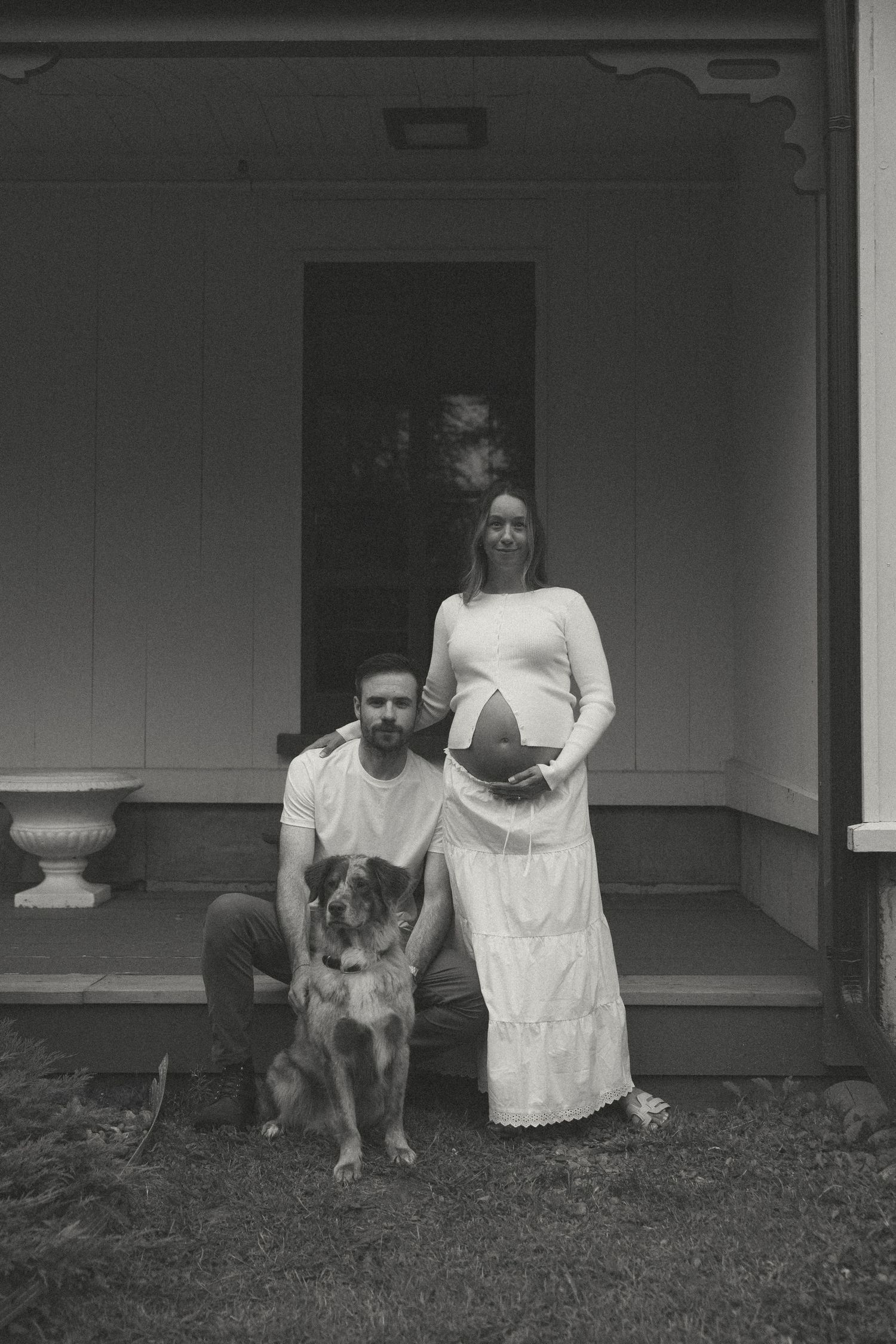 Black and white maternity photo of a couple with their dog on a porch, woman in white dress cradling baby bump.