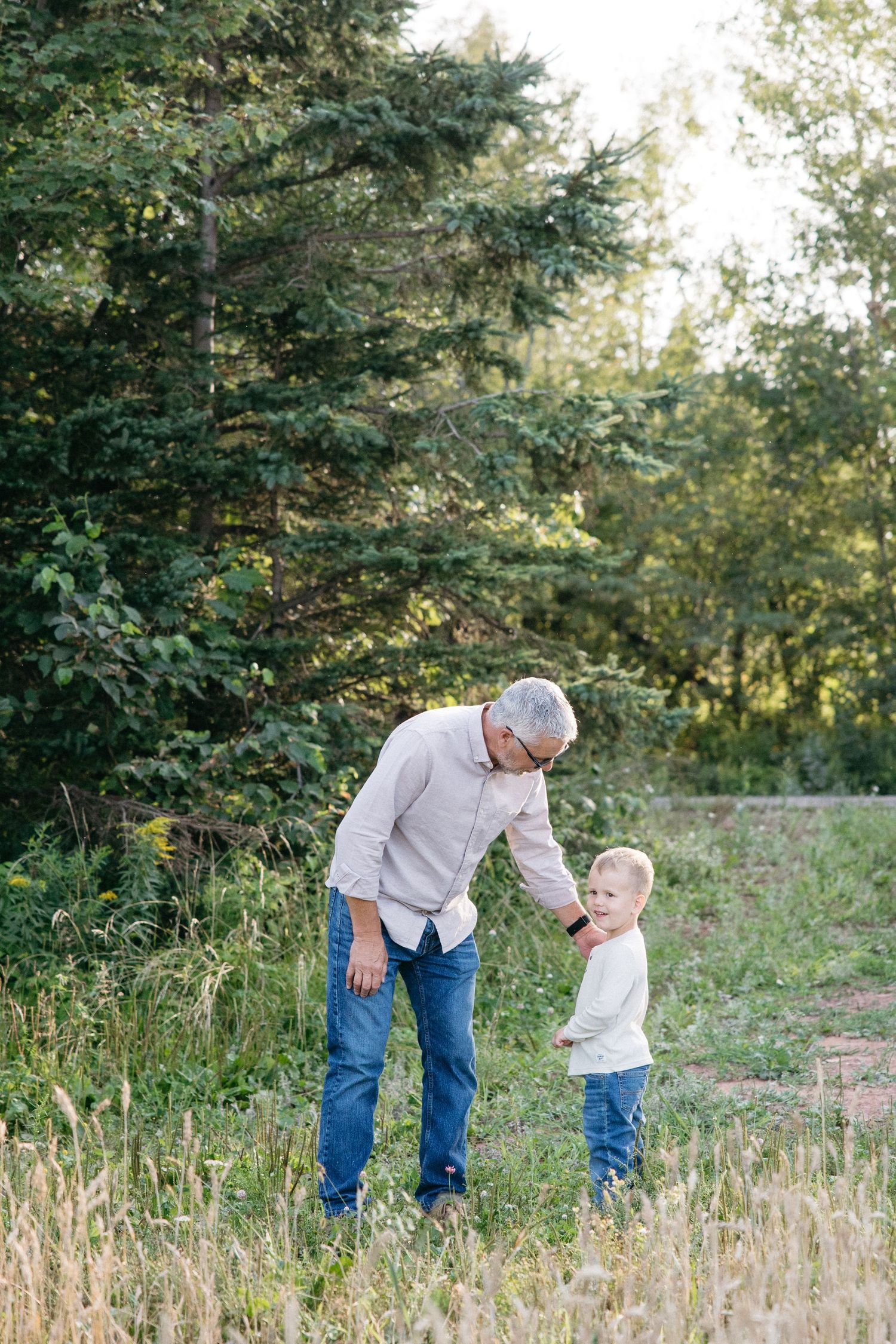 An older man bends down to interact with a young blonde toddler in a lush green outdoor setting.