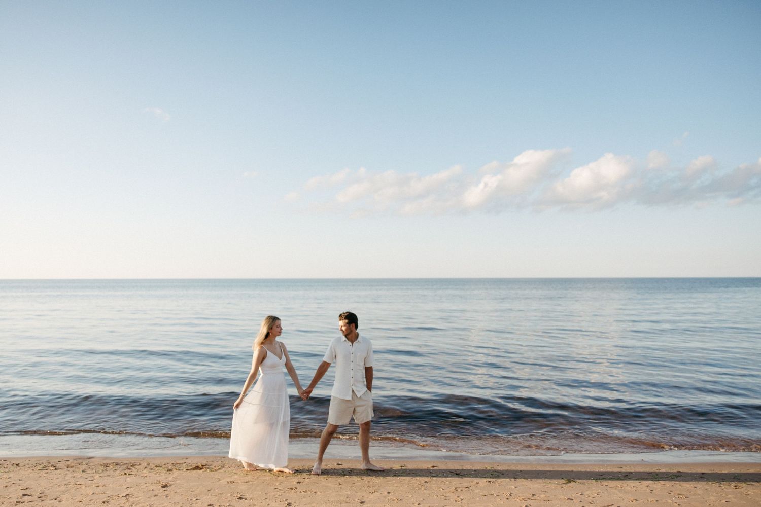 Couple in white attire holding hands on a sandy beach with calm blue ocean waters and clear sky backdrop.