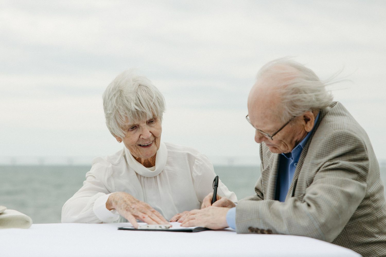 Elderly couple signing wedding documents together at an outdoor table with ocean view behind them.