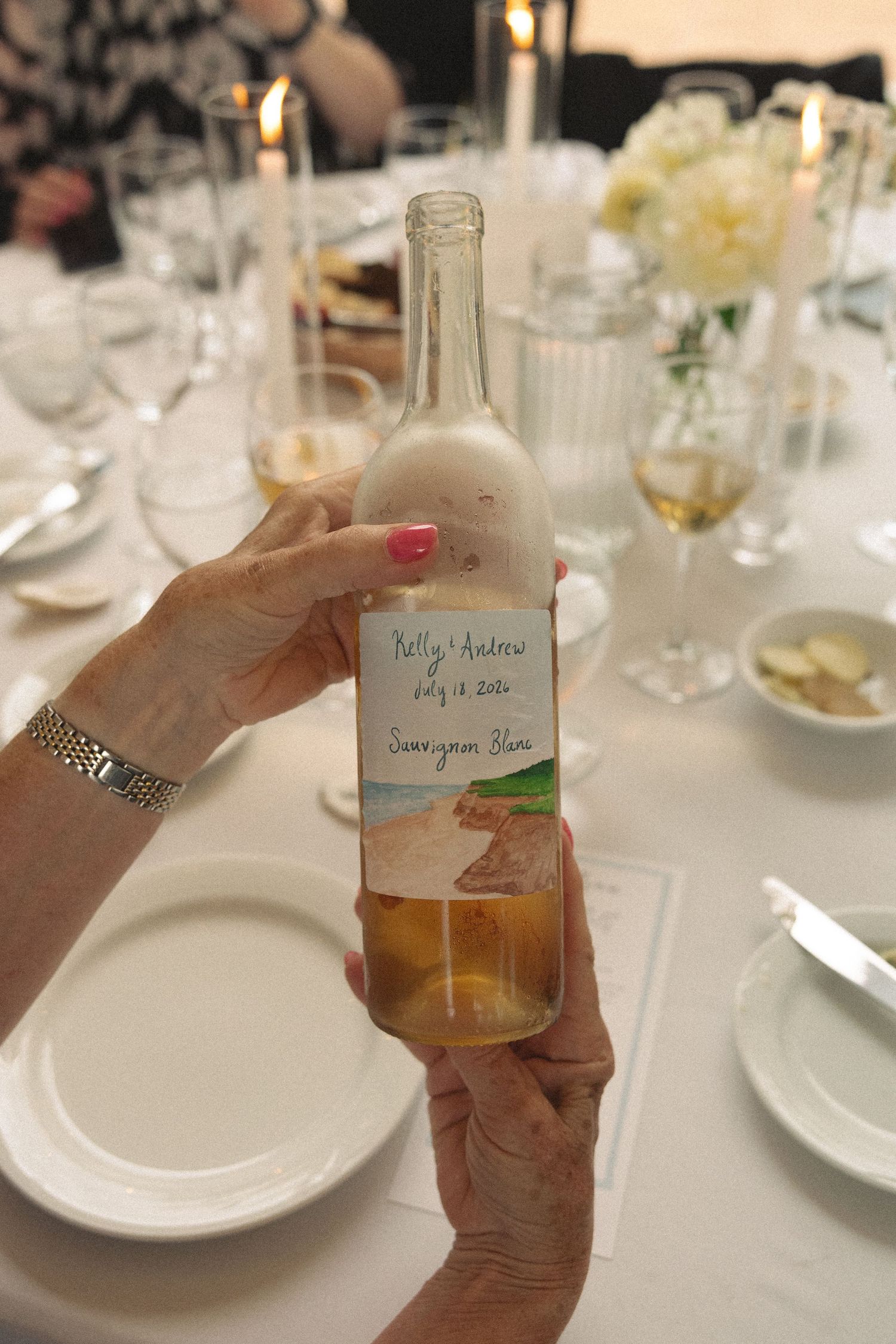 Hands holding a custom illustrated wedding wine bottle labeled Kelly and Andrew at an elegant reception table.