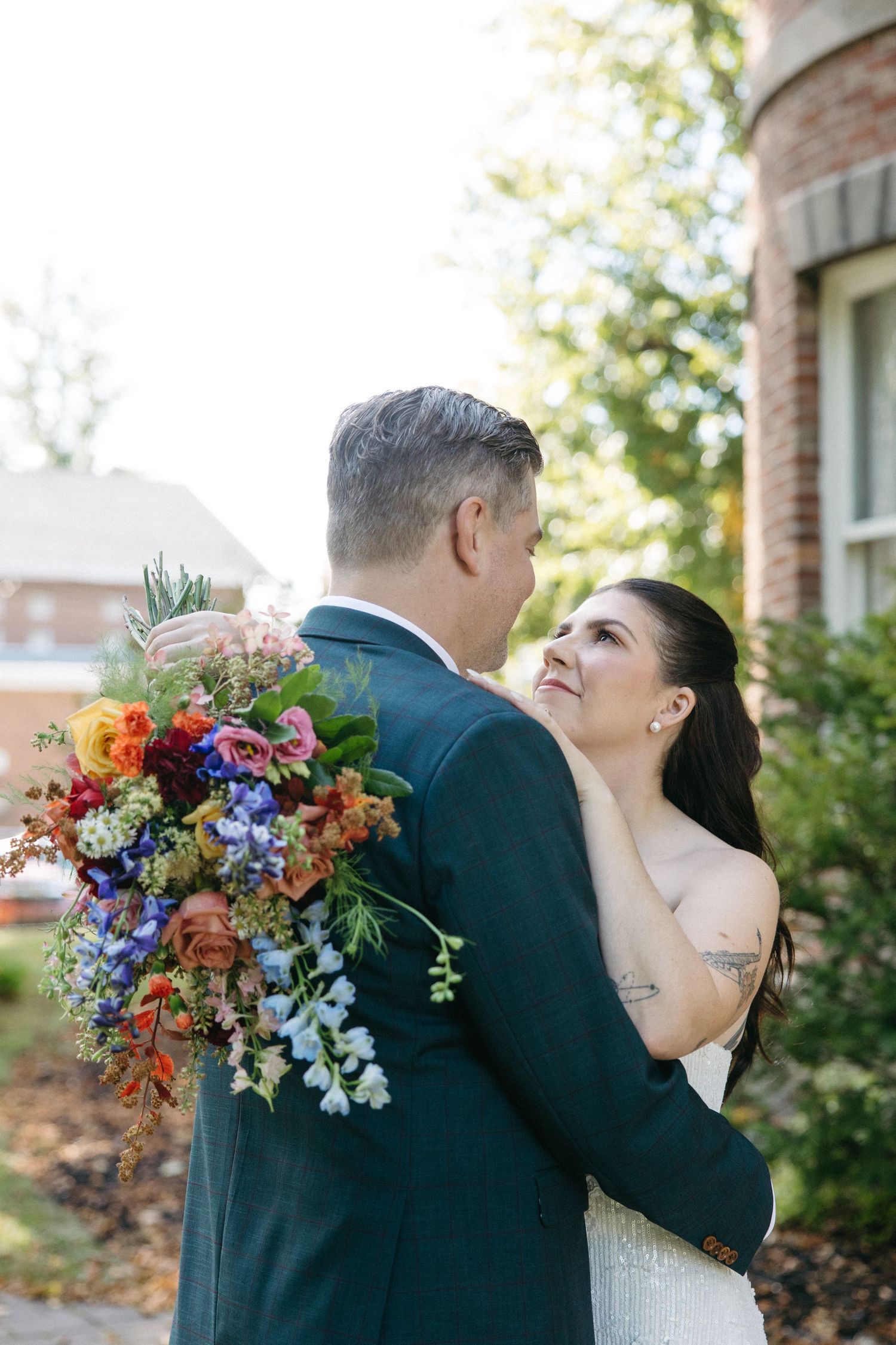 Bride in a white strapless gown embraces a groom in a dark suit while holding a colorful wildflower bouquet outdoors near a brick building.