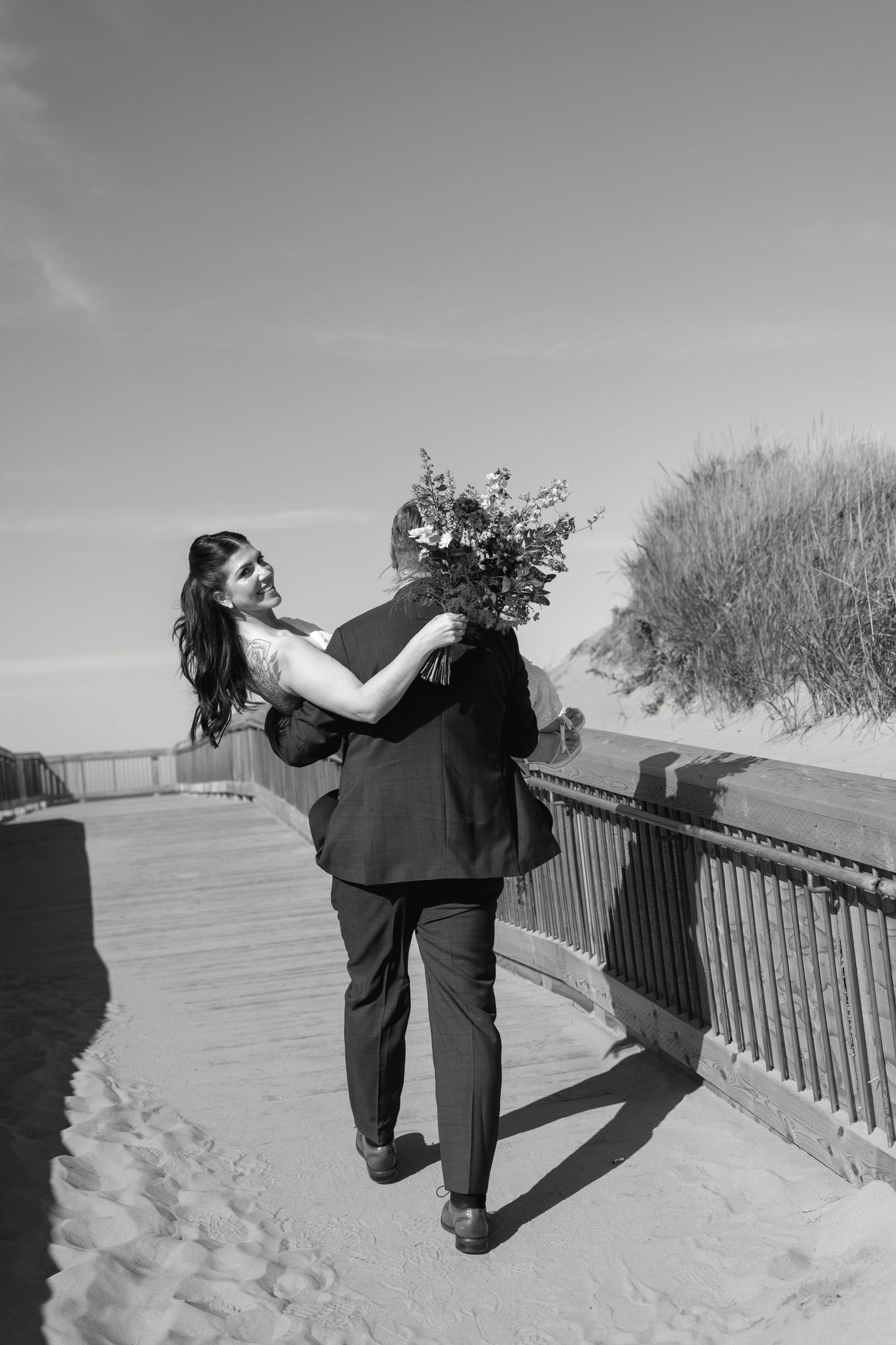 Black and white photo of a groom carrying a smiling bride holding a bouquet along a beach boardwalk beside sand dunes.
