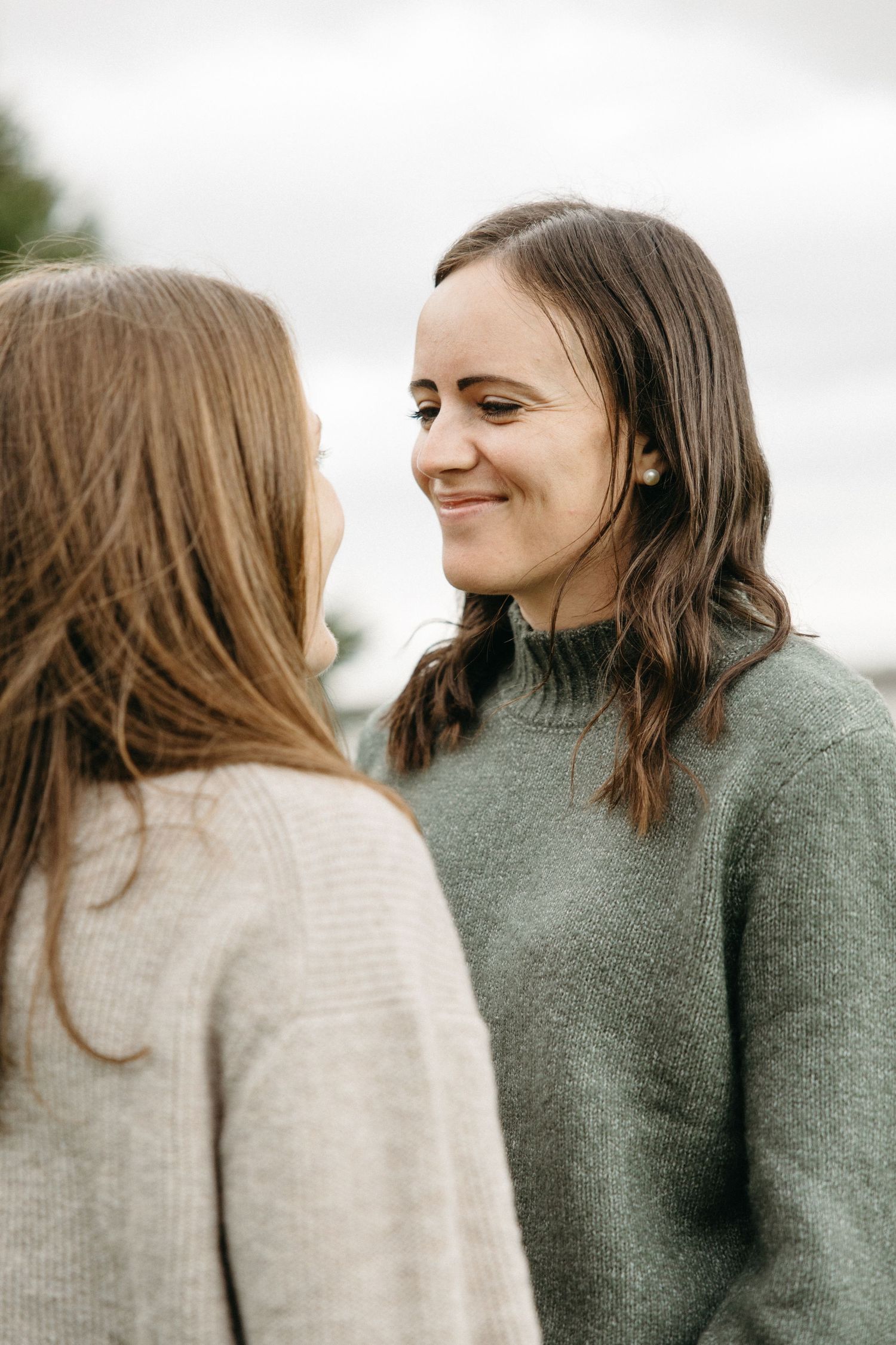 Two women stand face to face smiling warmly in cozy knit sweaters during an outdoor autumn engagement photo session.