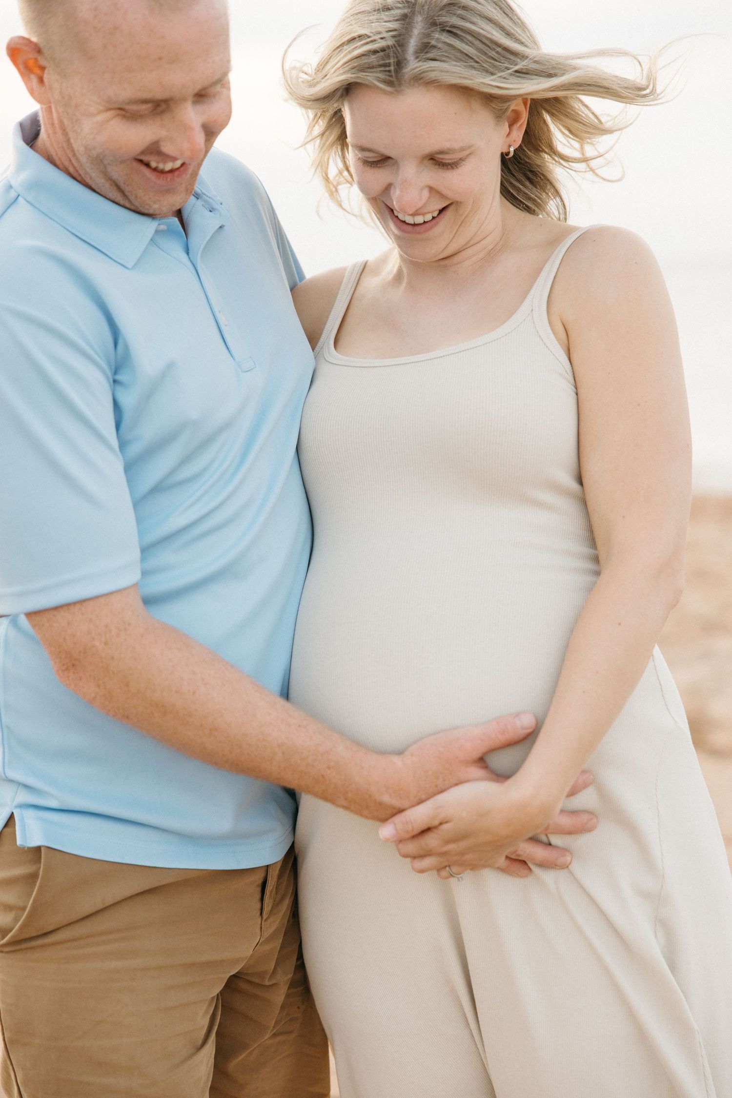 A smiling couple tenderly cradles a pregnant belly on a sunny beach, sharing a joyful maternity moment together.