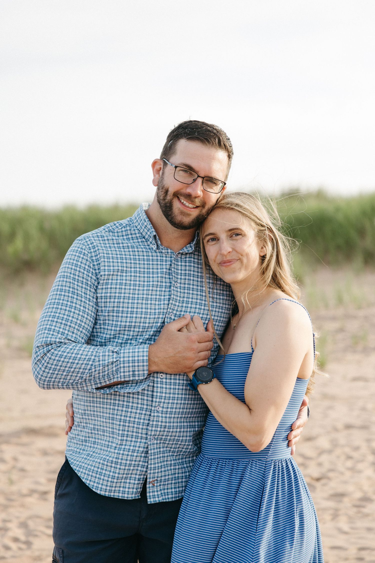 Happy couple embracing on a sandy beach, man wearing glasses and blue plaid shirt.