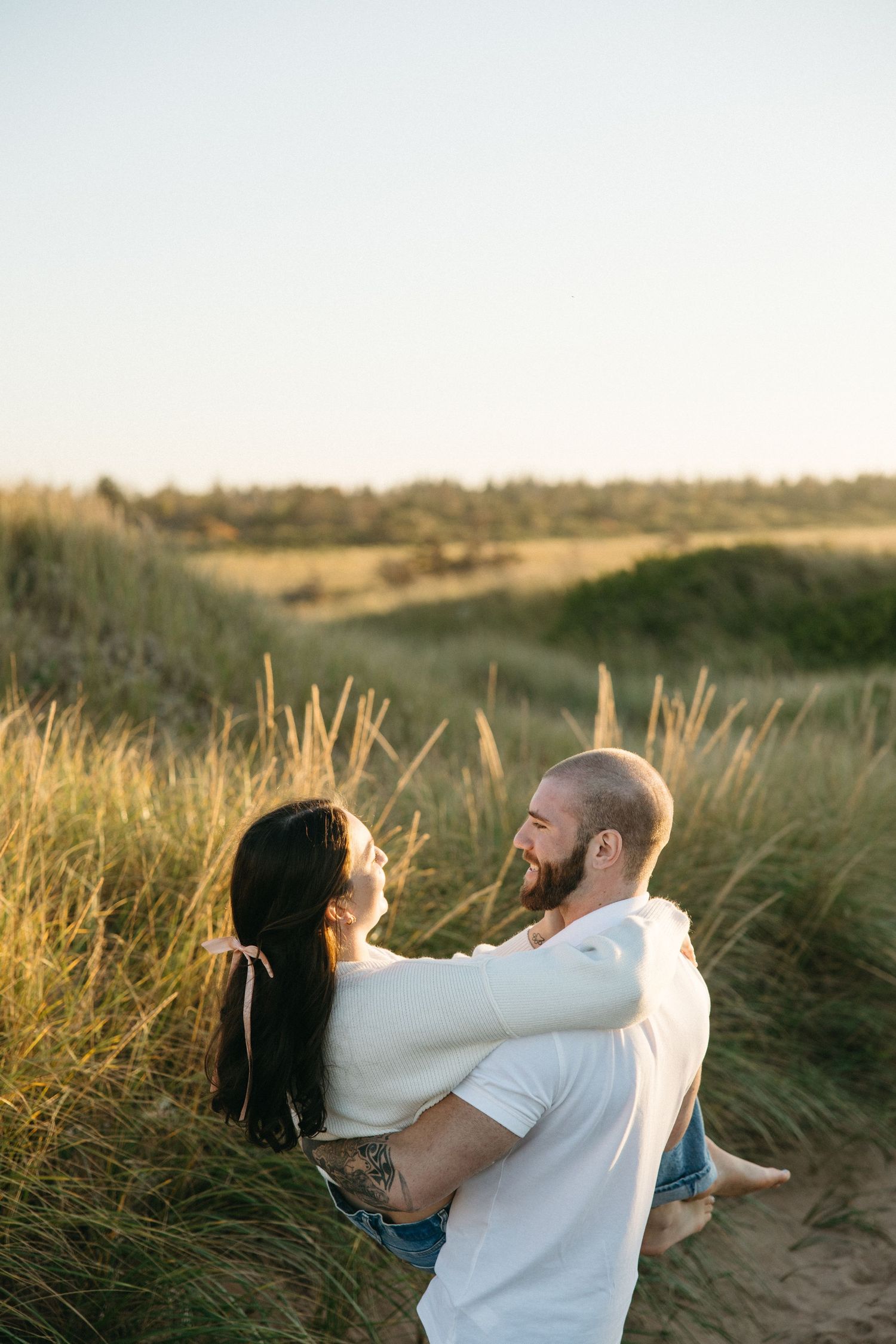 A couple embraces playfully in a field of tall grass during golden hour.