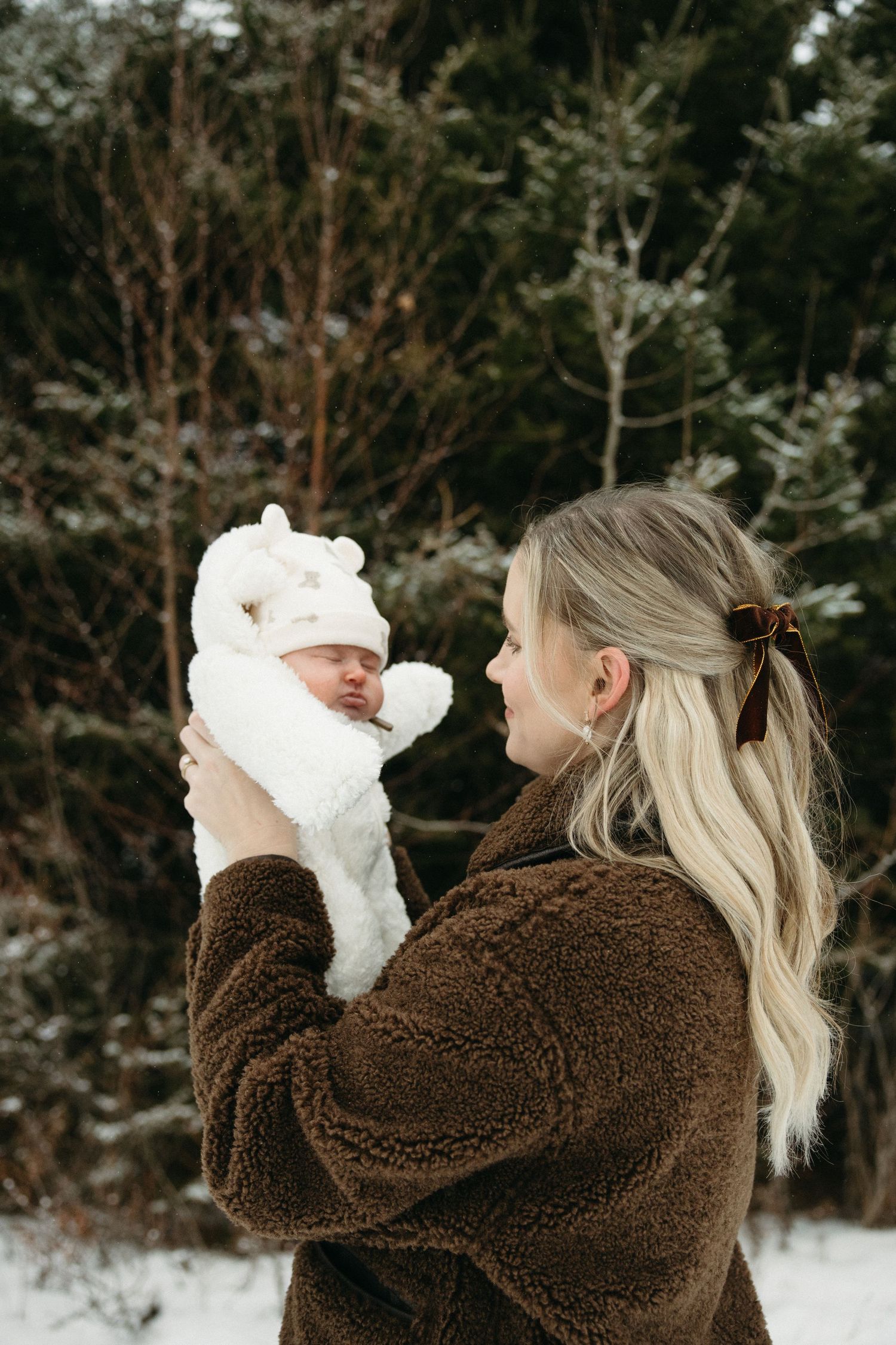 Mother in brown fleece jacket holding baby in white snowsuit against snowy forest backdrop.