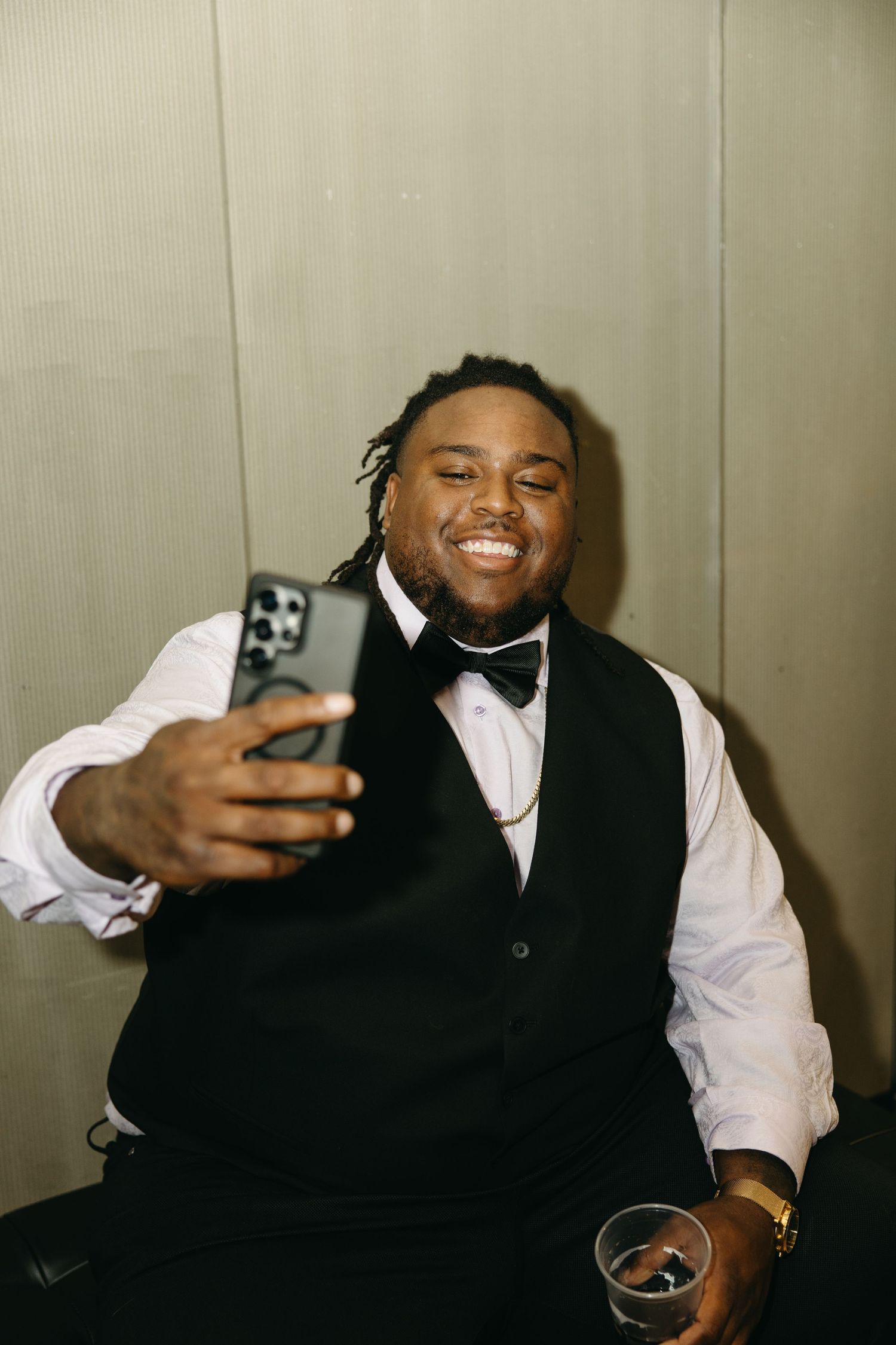 Person in formal attire taking selfies and smiling while holding a drink.
