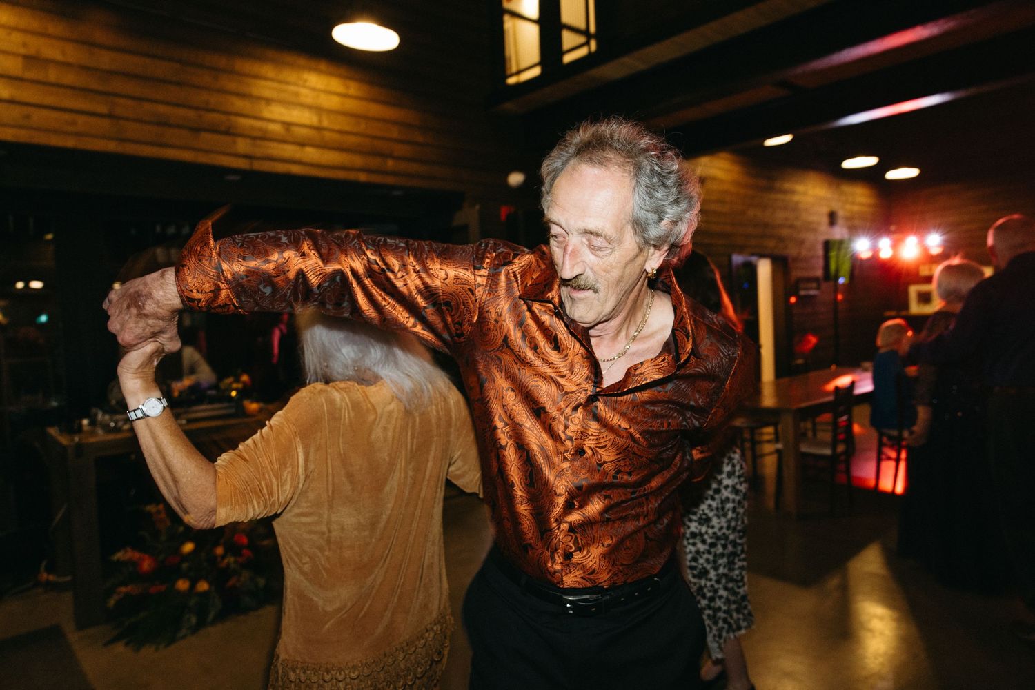 Wedding guests dance and celebrate in a dimly lit venue with wooden walls and ceiling.