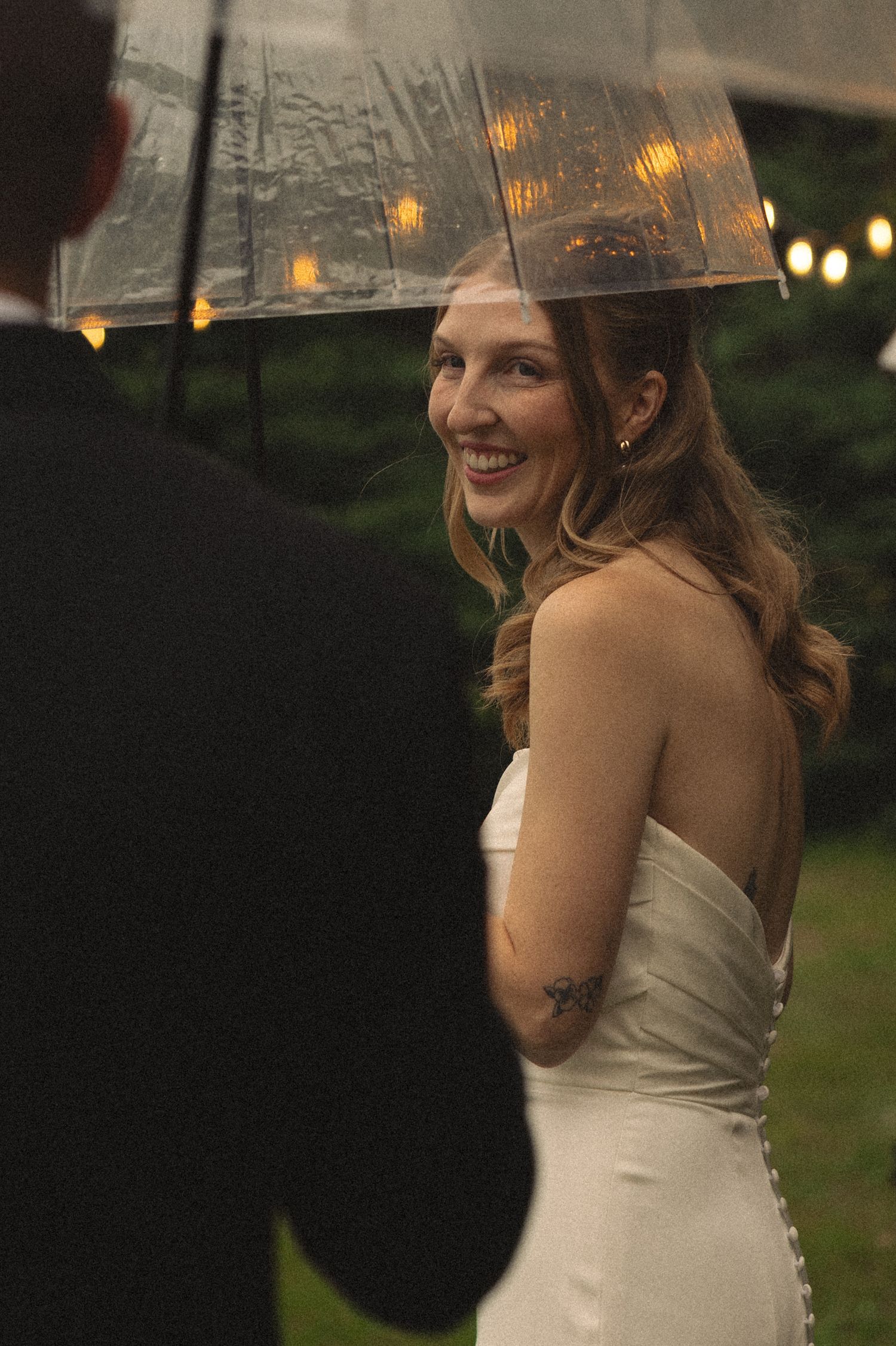 Smiling bride in strapless white gown holds clear umbrella with string lights reflected, sharing romantic rainy outdoor wedding moment.