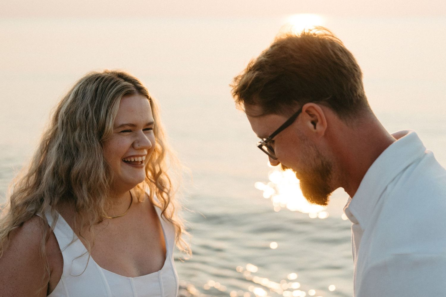 Couple laughing together at sunset by the ocean, woman in white dress with golden backlight.
