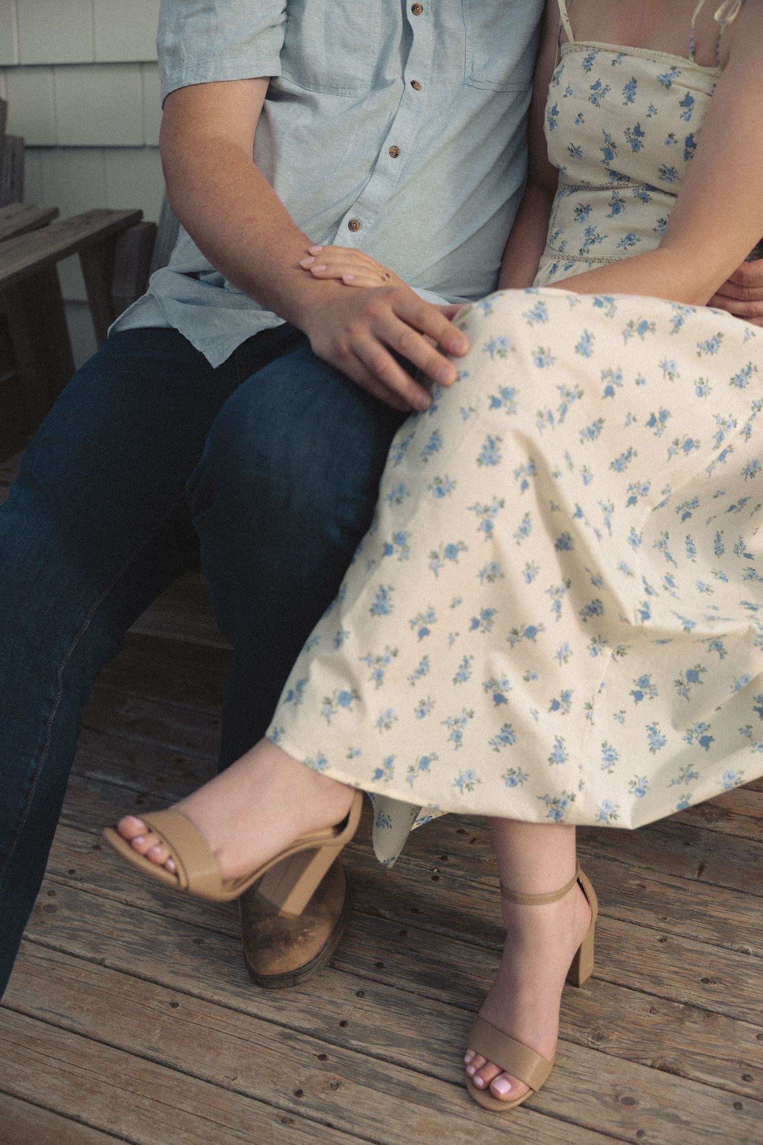 Close-up of a couple holding hands while sitting together, woman wearing a floral midi dress and tan heels.