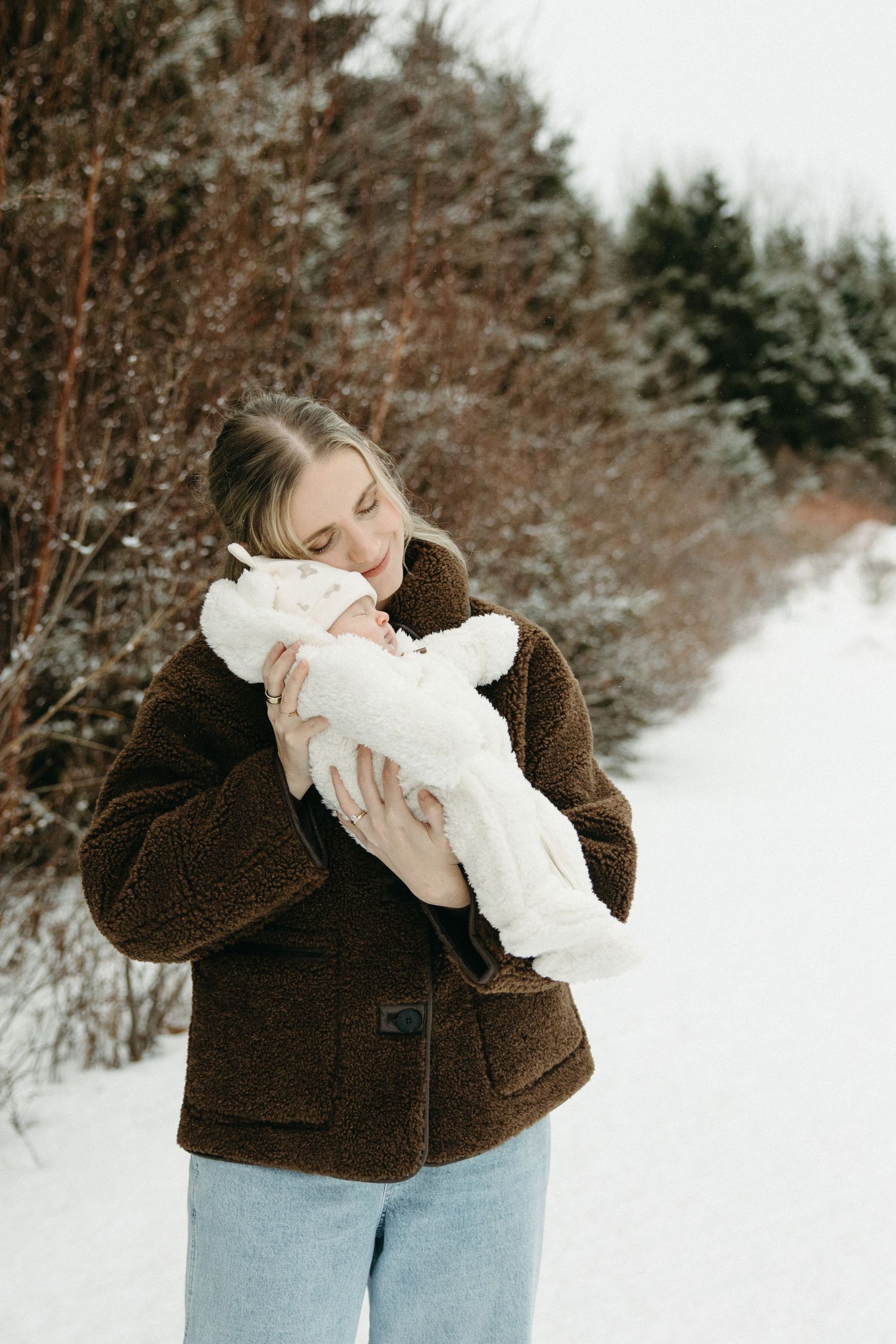 Woman in brown jacket holding baby dressed in white against snowy landscape.