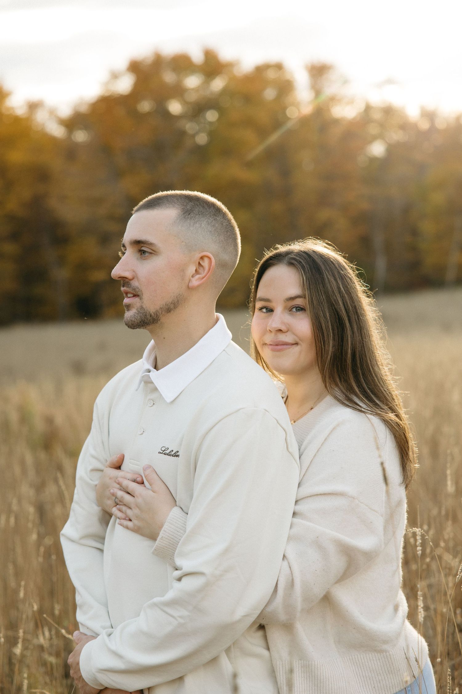 A couple in cream-colored sweaters embraces in a sunlit autumn field.