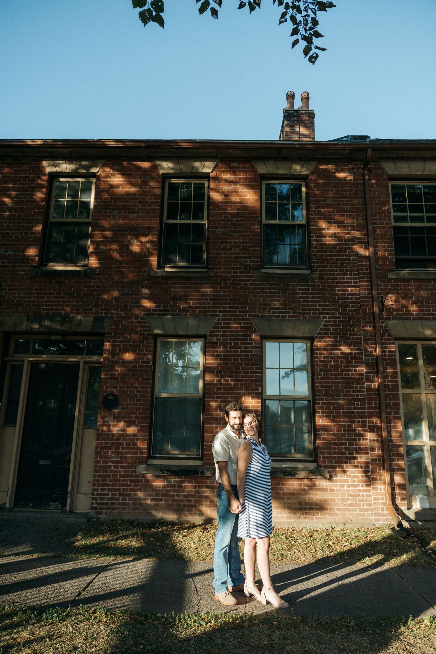 A romantic couple poses in front of a vintage brick building with dappled sunlight.