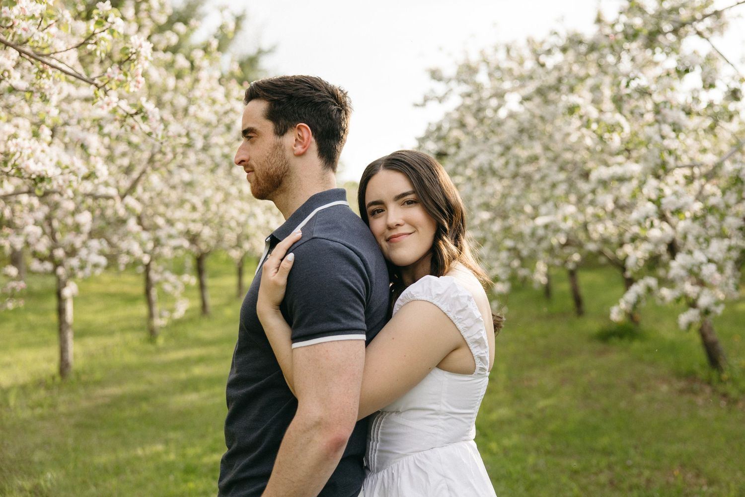 A woman in a white dress hugs a man from behind while smiling in a blossoming orchard.