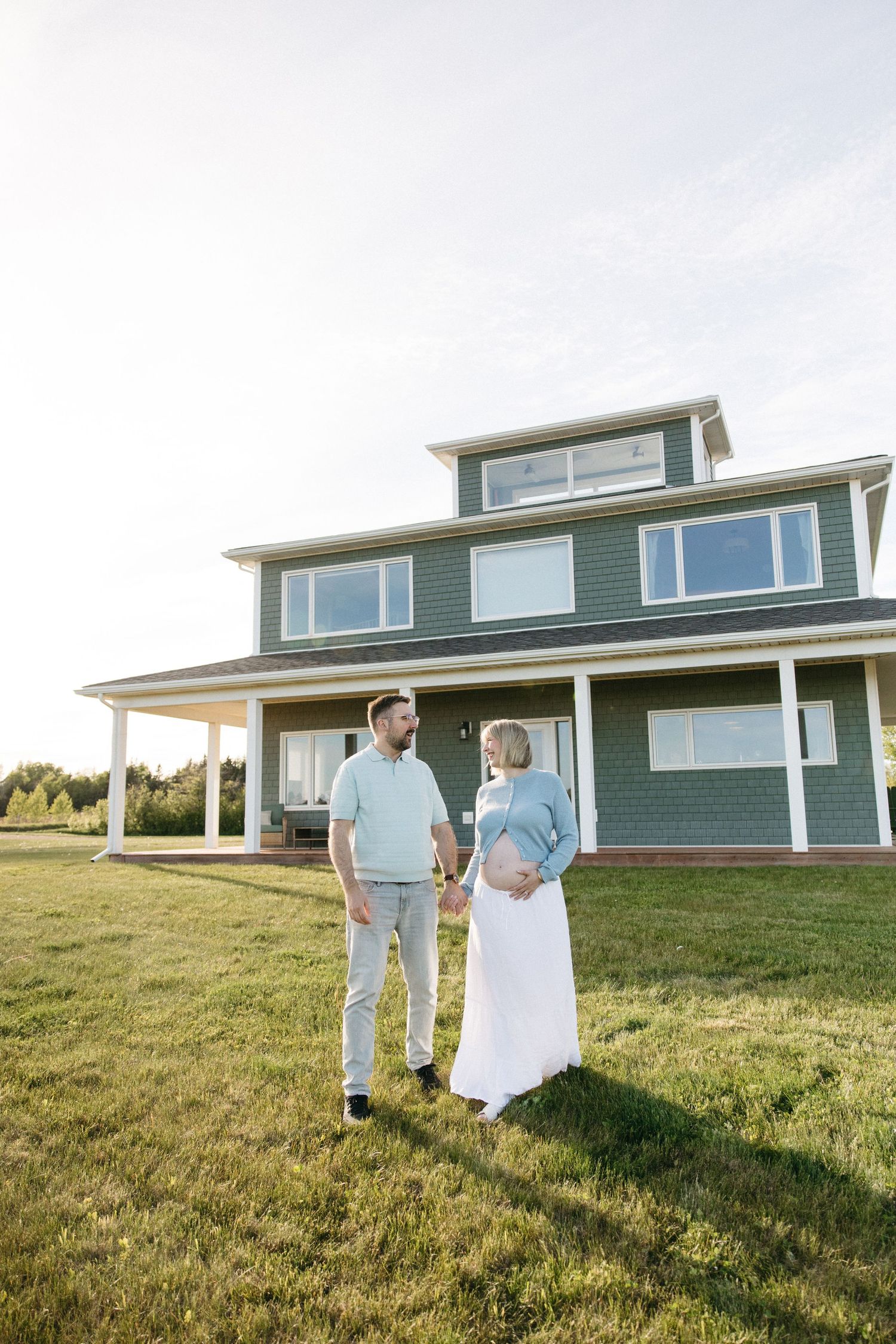 Expecting couple holds hands in front of large green two-story home on sunny day.