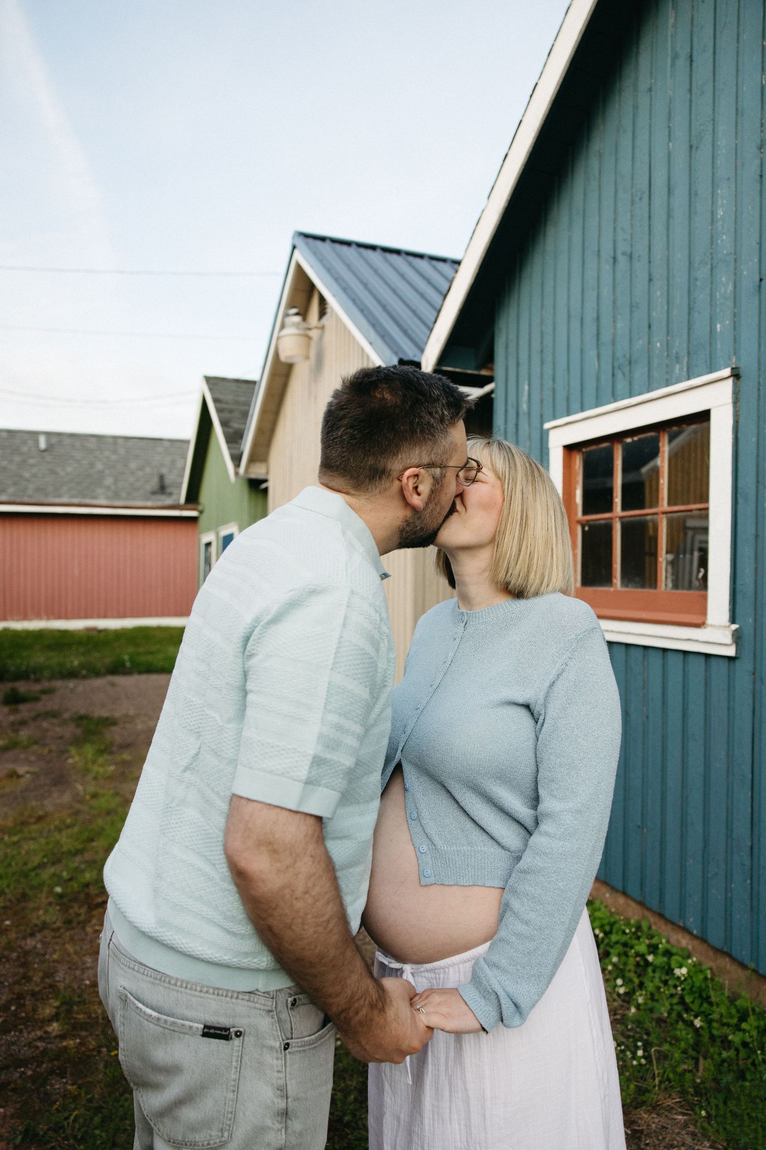 Couple kissing outdoors near colorful barn buildings, pregnant woman in blue sweater.