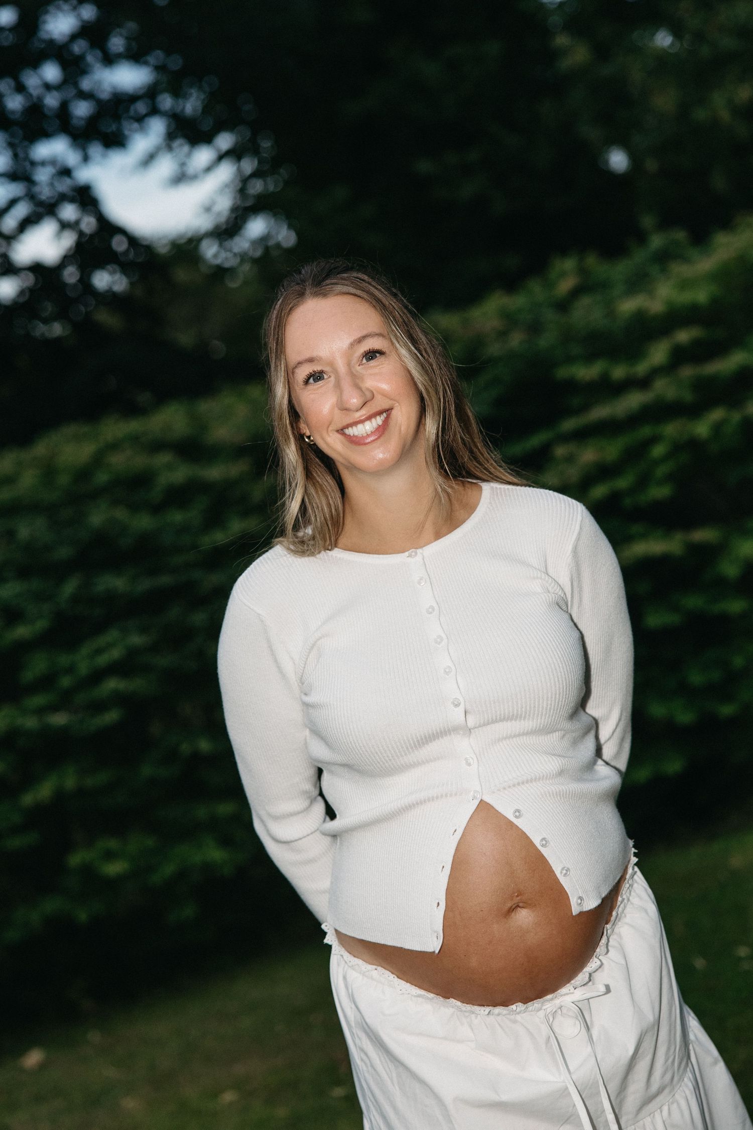 Smiling pregnant woman in white outfit showing baby bump outdoors in lush green garden setting.