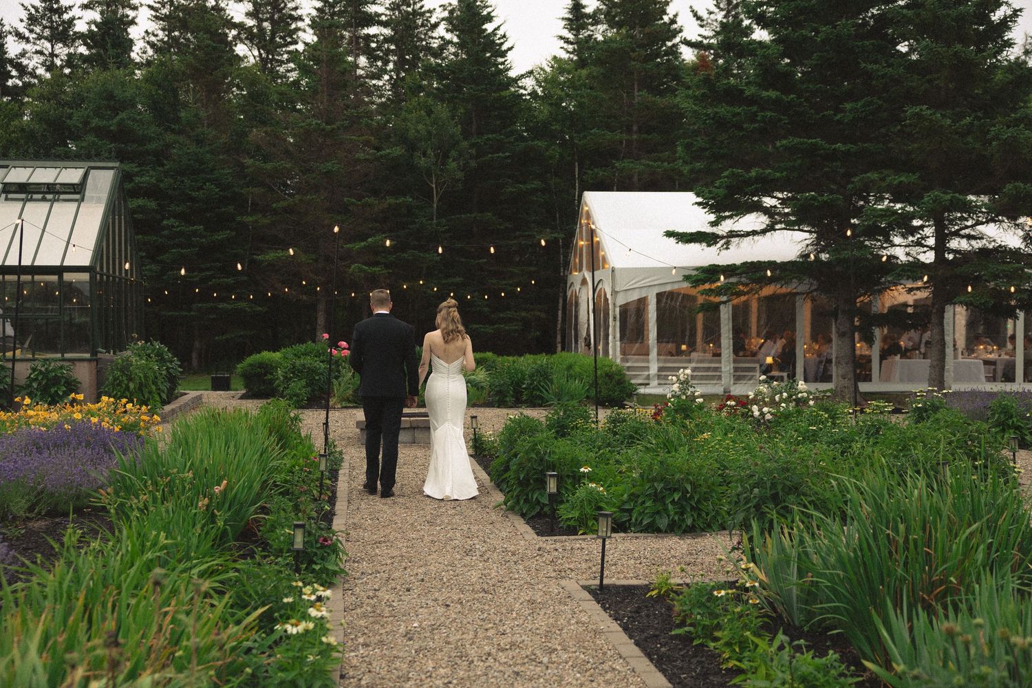 Bride and groom walking hand in hand through garden path toward lit reception tent at dusk.