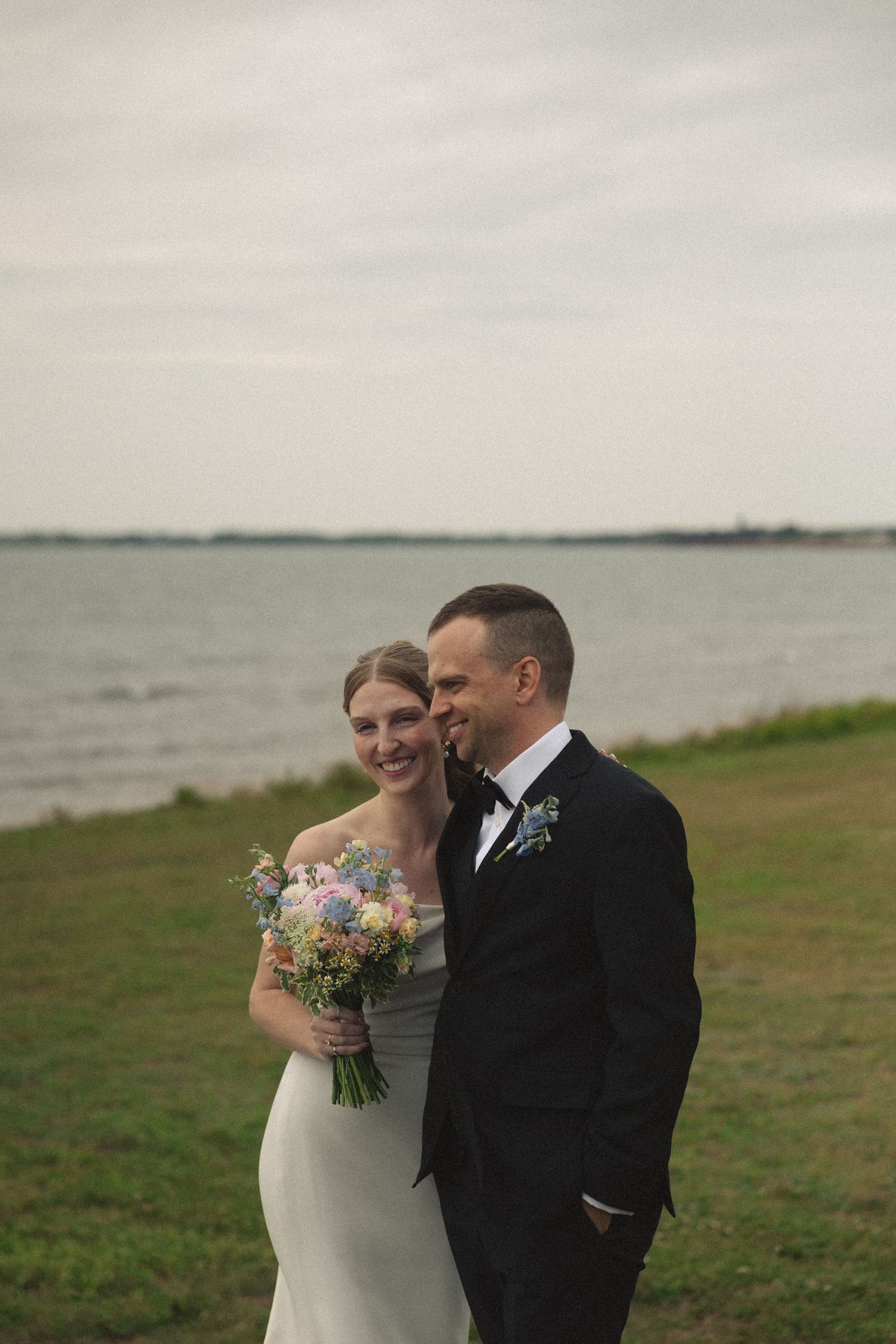 Bride and groom pose romantically by waterfront, she holds a pastel wildflower bouquet in a white gown.