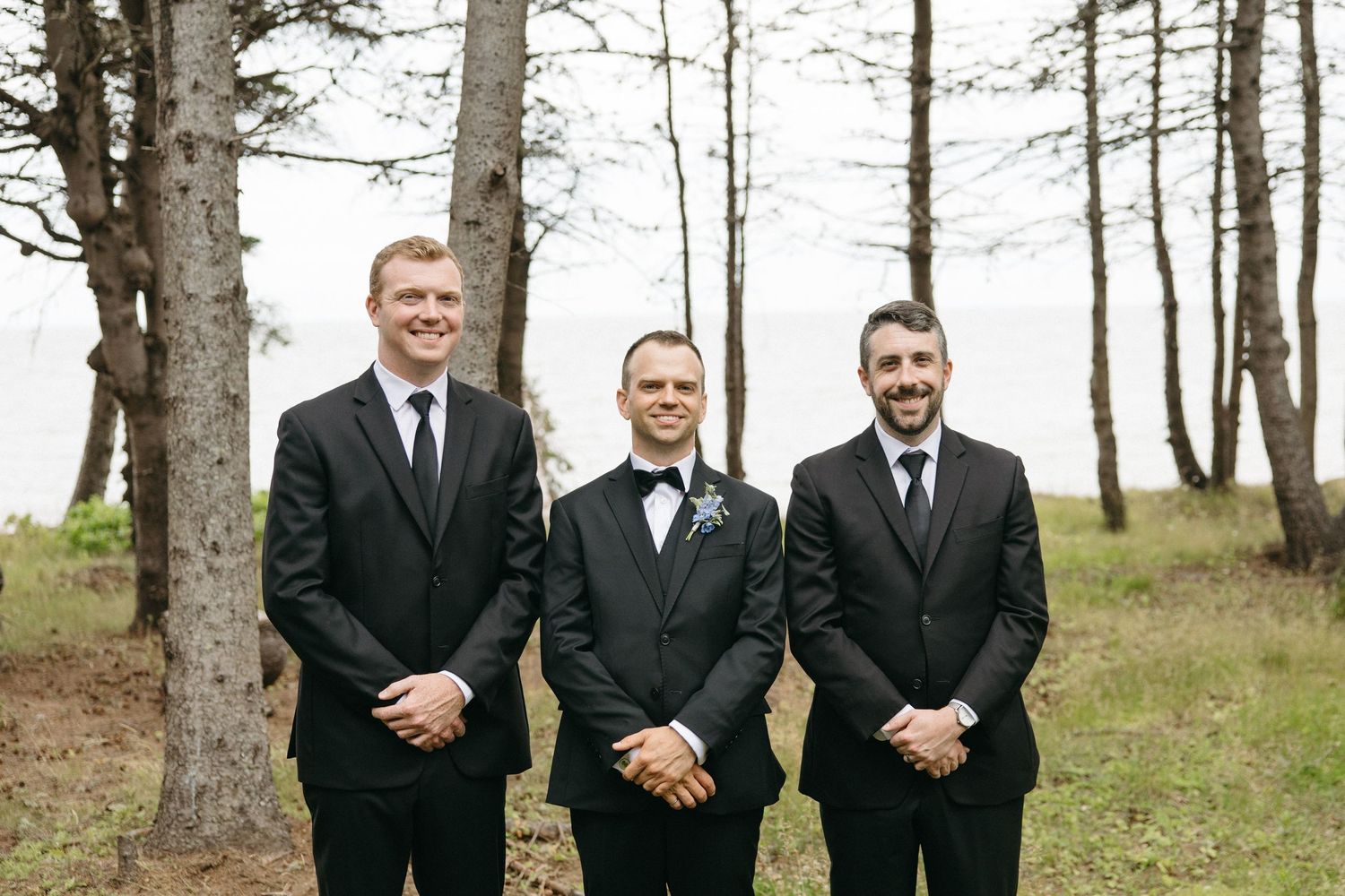 Groom and two groomsmen in black suits smiling outdoors among tall trees.