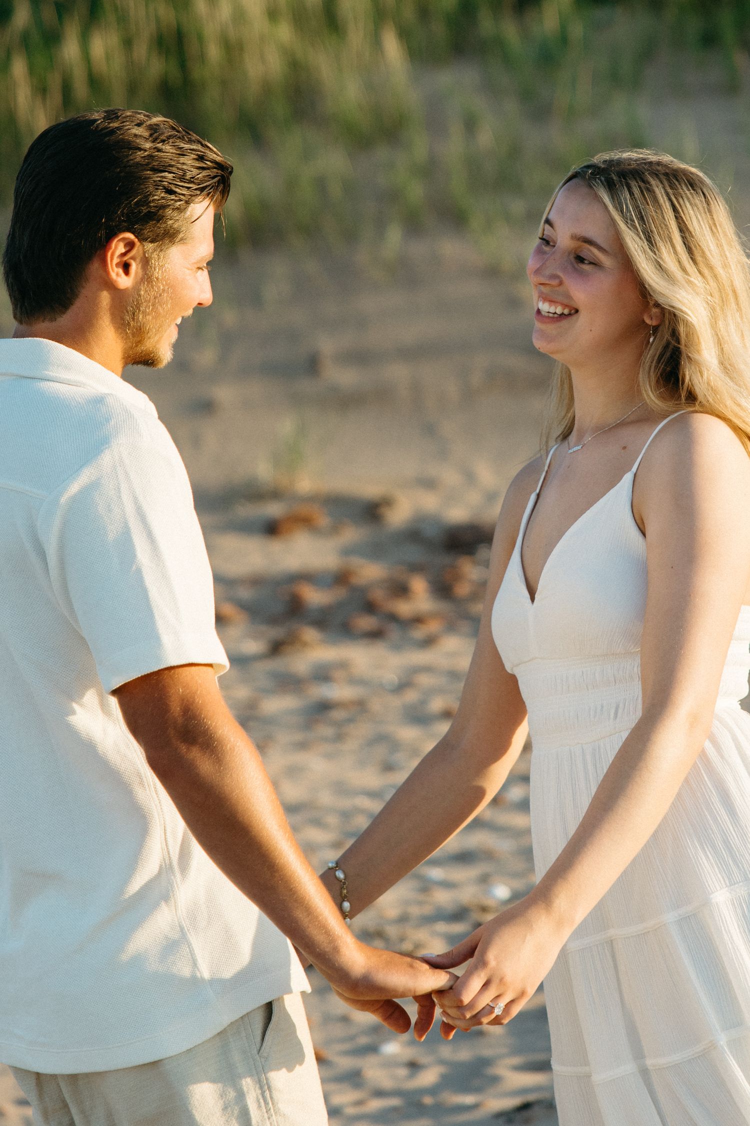 Happy blonde woman in white dress holding hands with man on sandy beach in golden sunlight.