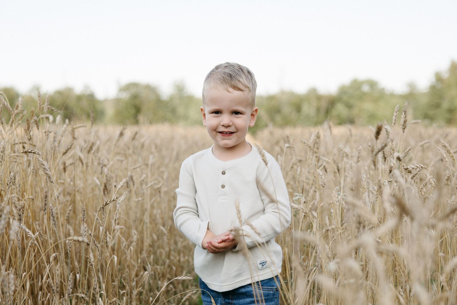 Young boy smiling in a golden wheat field wearing a white henley shirt and jeans on a bright day.