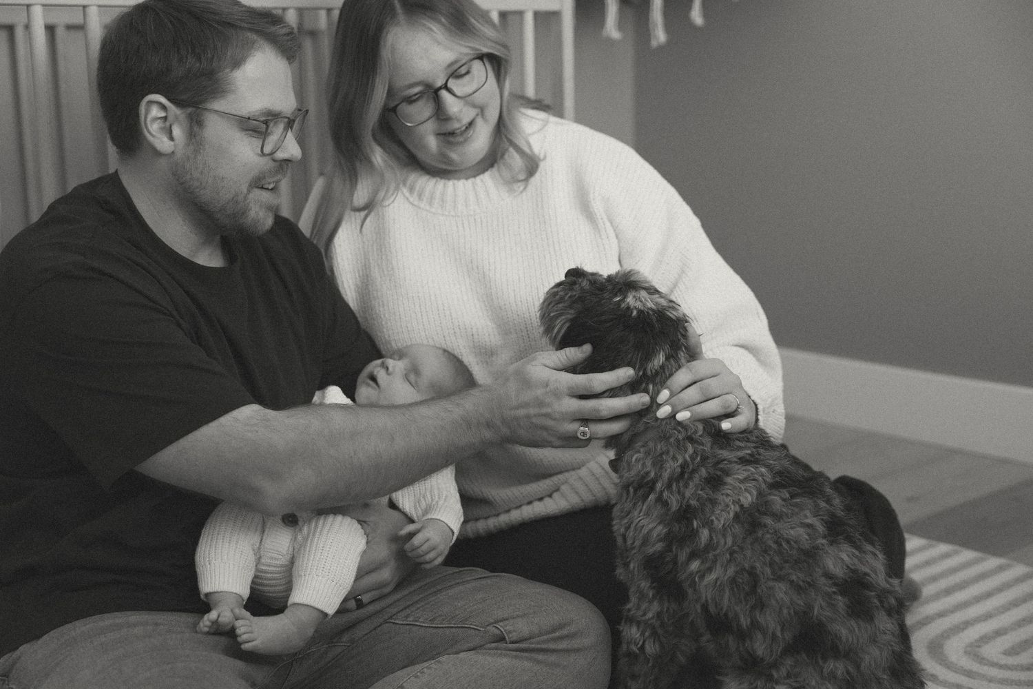A couple sits on a couch sharing a tender moment with their small black dog in a black and white photograph.