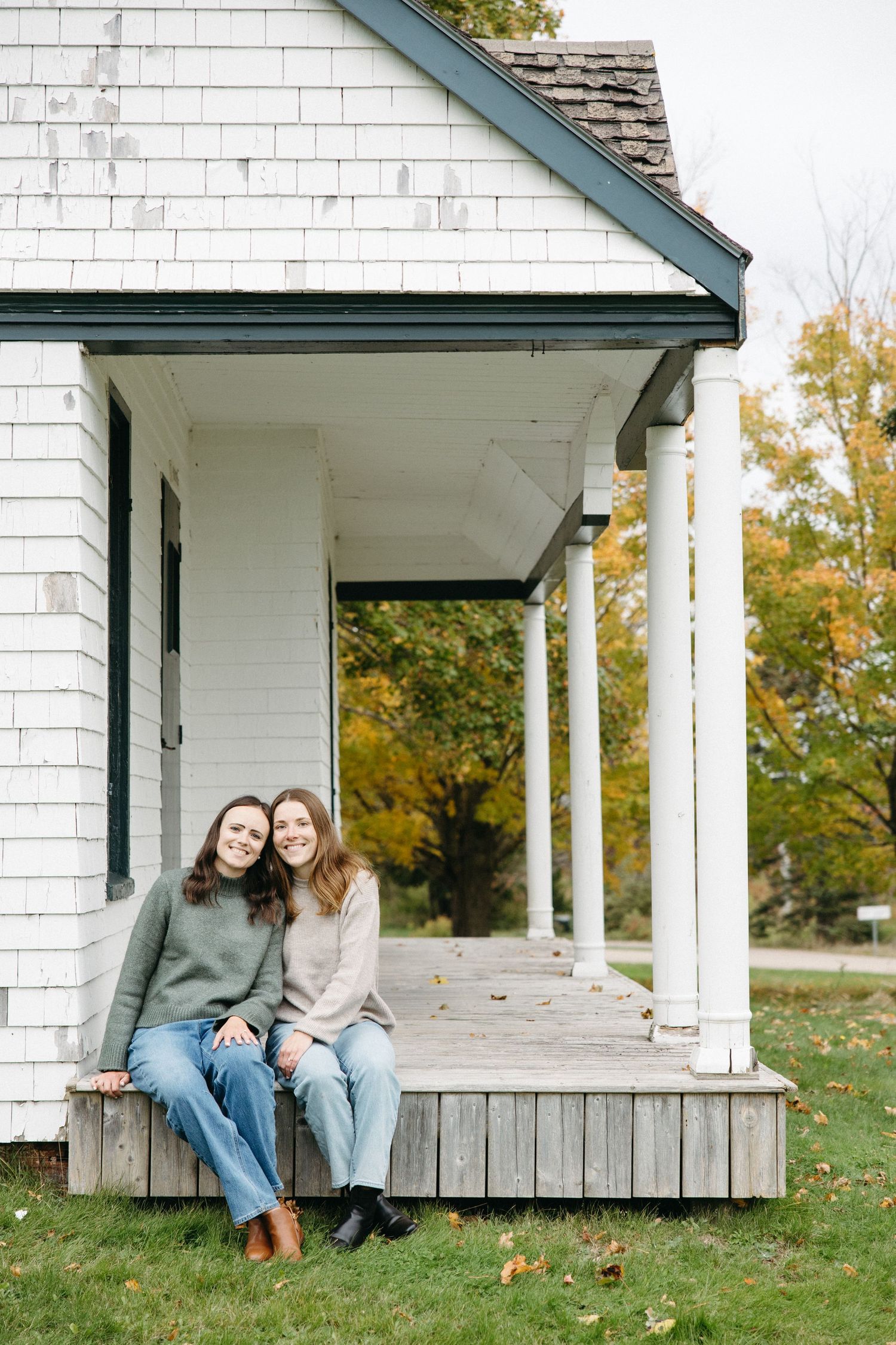 Two women in sweaters and jeans sit smiling on the edge of a wooden porch of a white clapboard house surrounded by autumn trees.