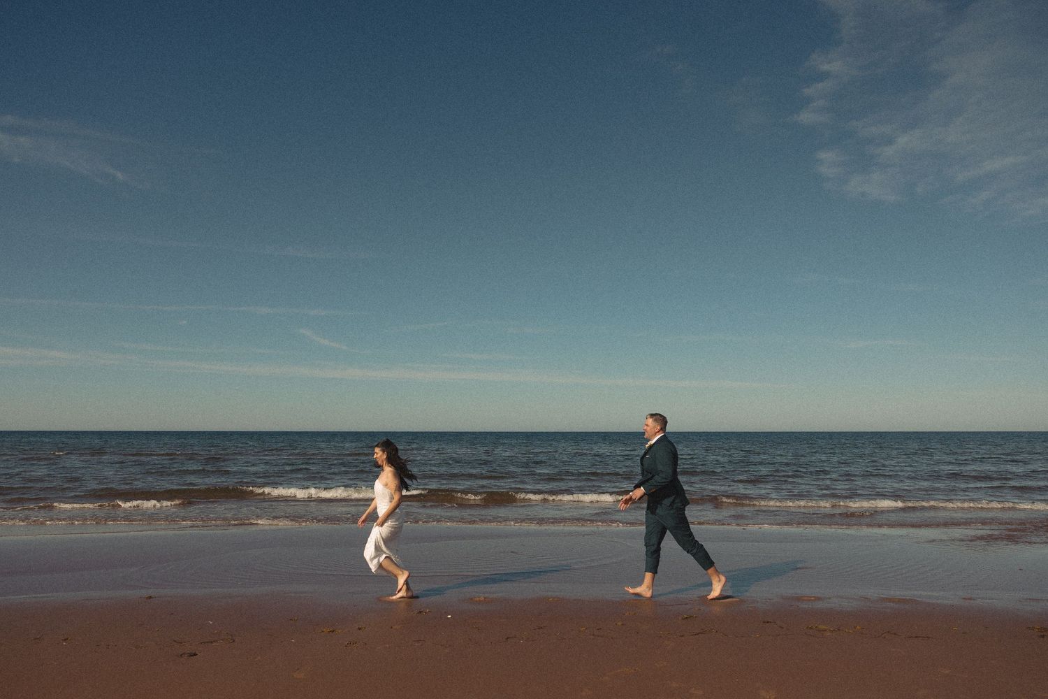 Couple running barefoot along the wet sand of a beach toward gentle ocean waves under a blue sky with wispy clouds.