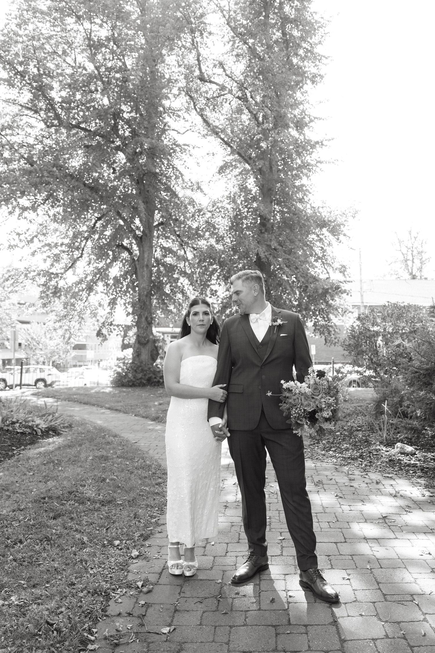 Black and white photo of a bride in a white dress and groom in a dark suit holding a bouquet, walking arm in arm on a brick path.