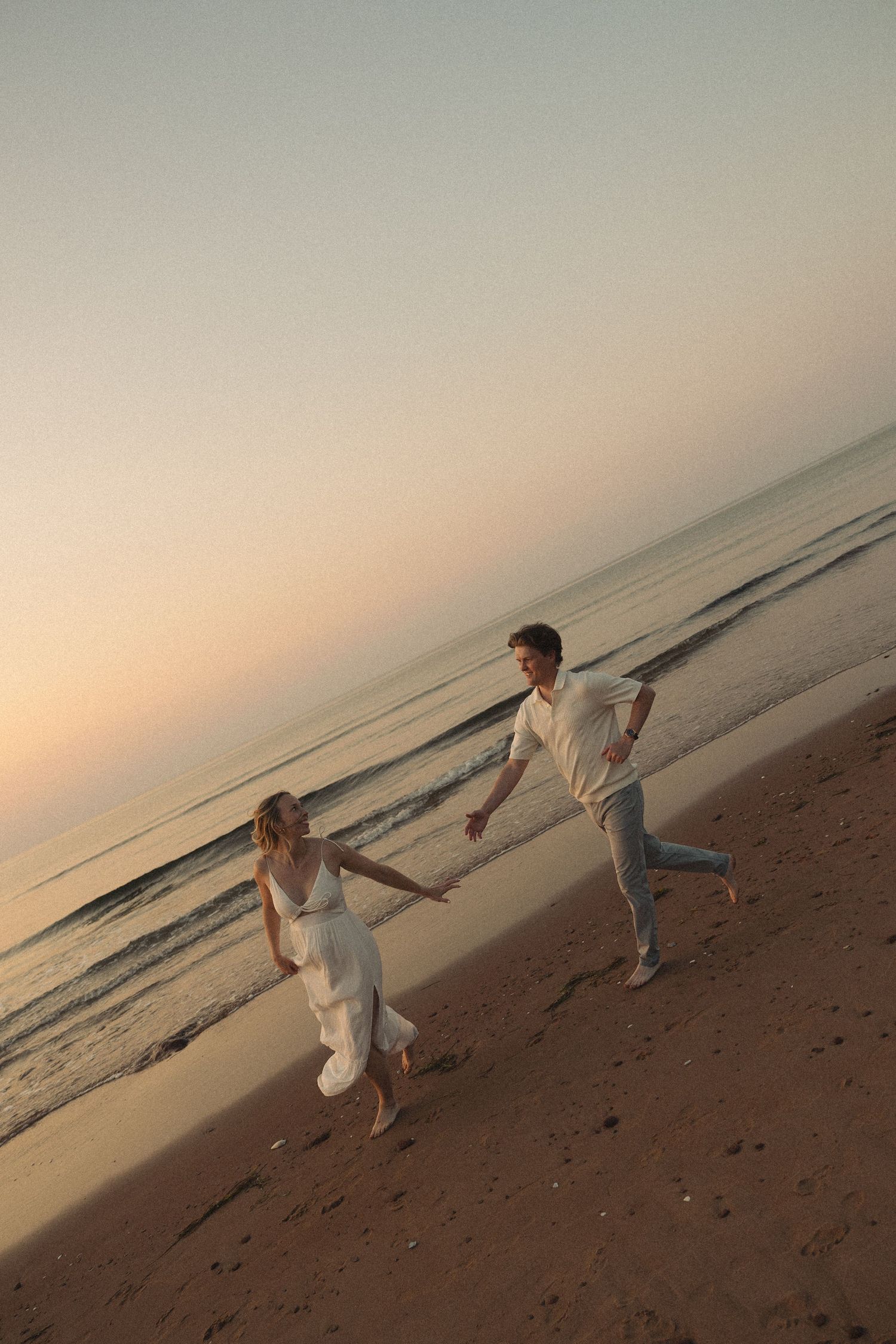 Couple holding hands and running along sandy beach at dusk with serene ocean backdrop.