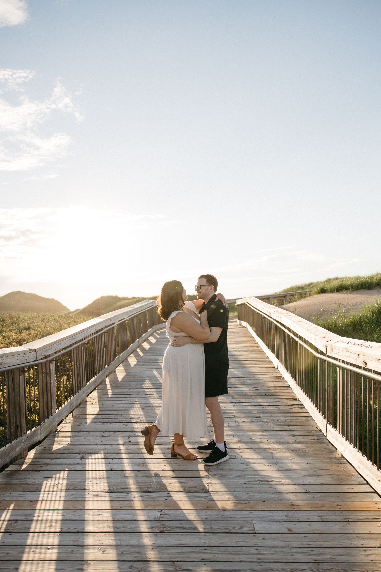 A couple embraces and kisses on a wooden boardwalk overlooking scenic green hills at golden sunset.