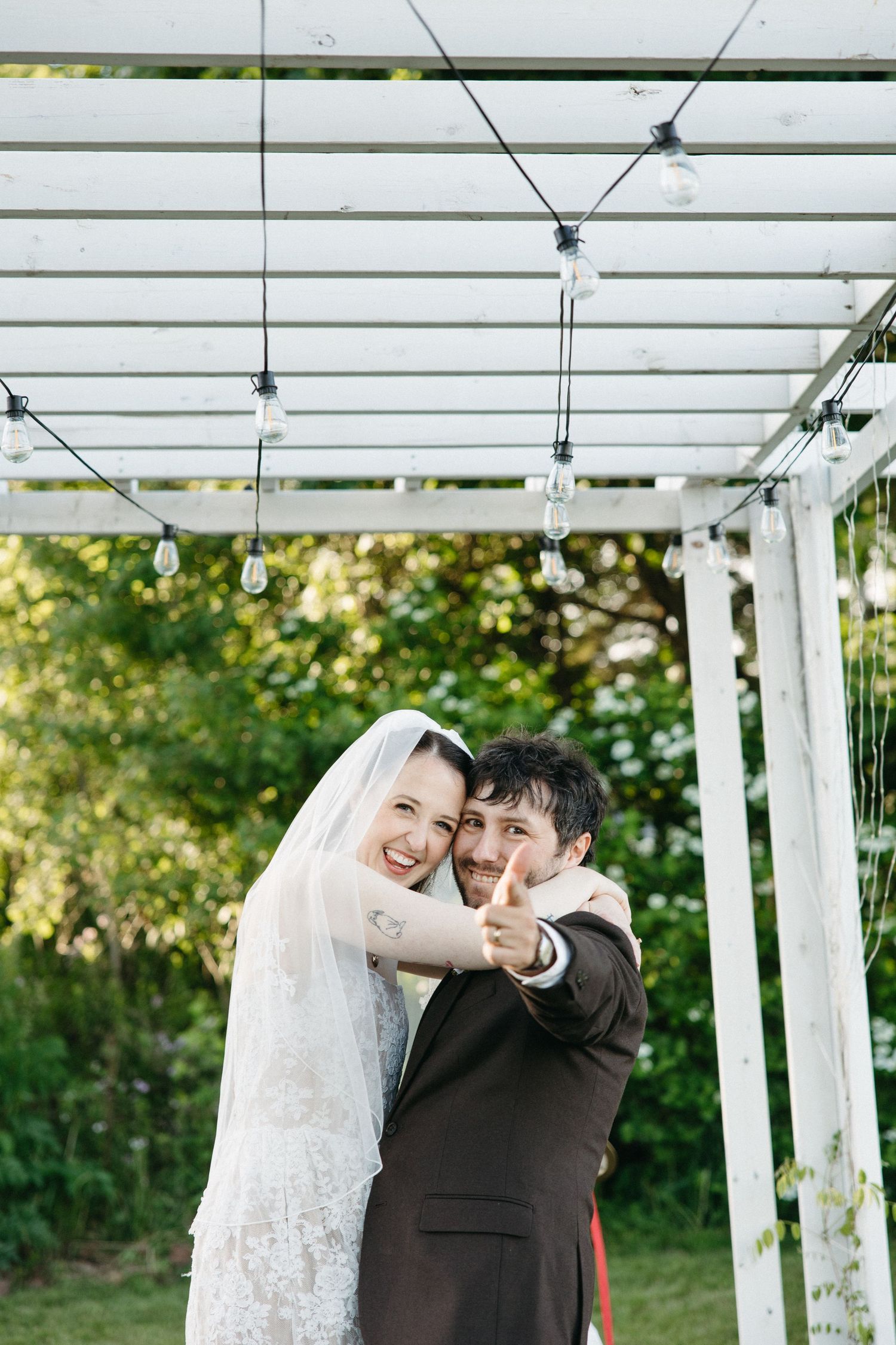 Smiling bride and groom hugging outdoors under white pergola with string lights, groom giving thumbs up on wedding day.