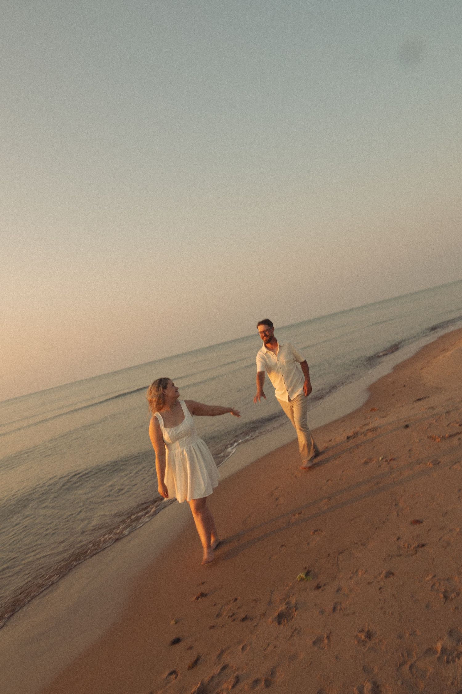 Couple in white outfits holding hands while walking along a sandy beach at golden sunset.