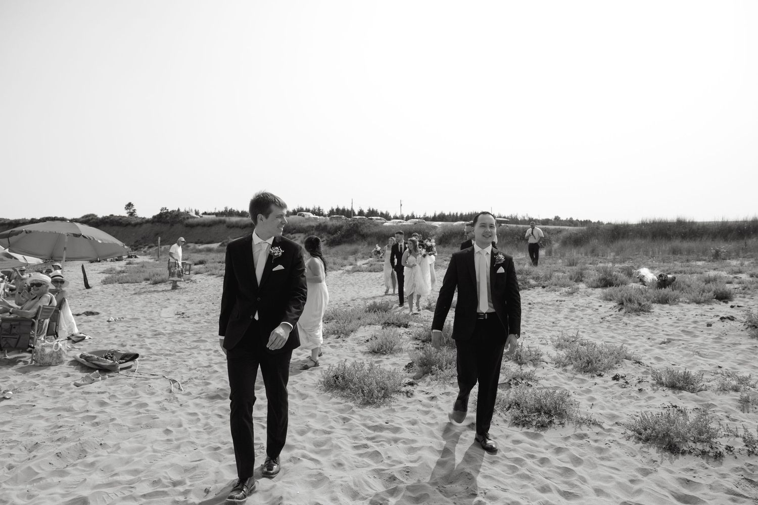 Two figures in dark suits walk across a sandy beach landscape in black and white.