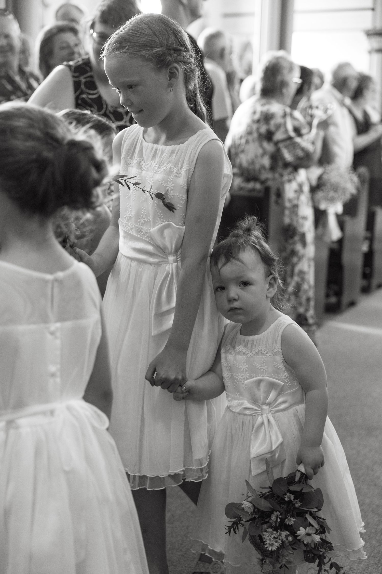 Young flower girls in white dresses holding bouquets line up during a wedding ceremony.